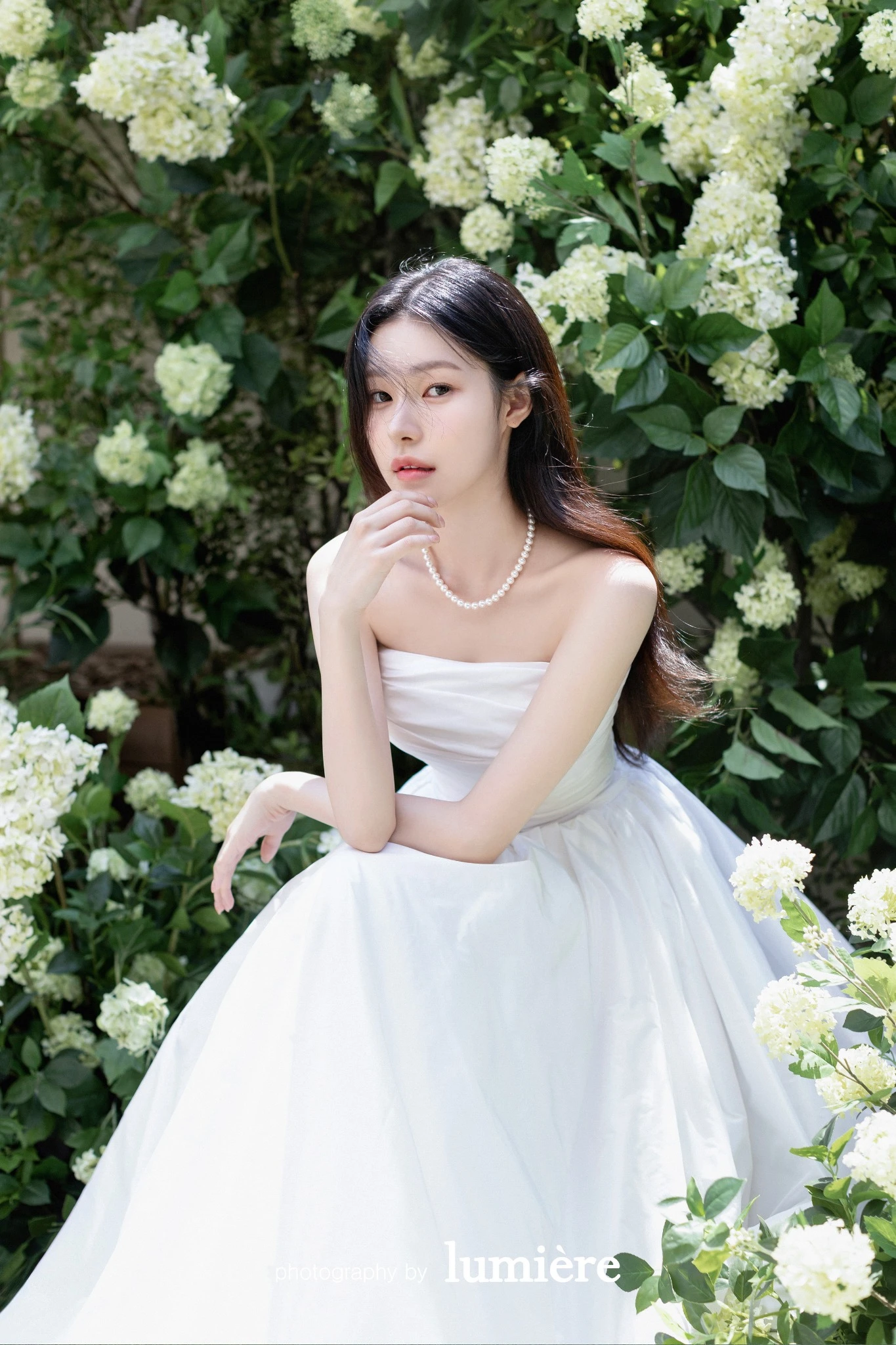 흰 꽃이 많은 그리너리 배경에서 웨딩드레스를 입은 여성 단독이 앉은 자세로 손을 턱 근처에 둠 모습이다. 흰 꽃·진주 목걸이가 함께 보인다.