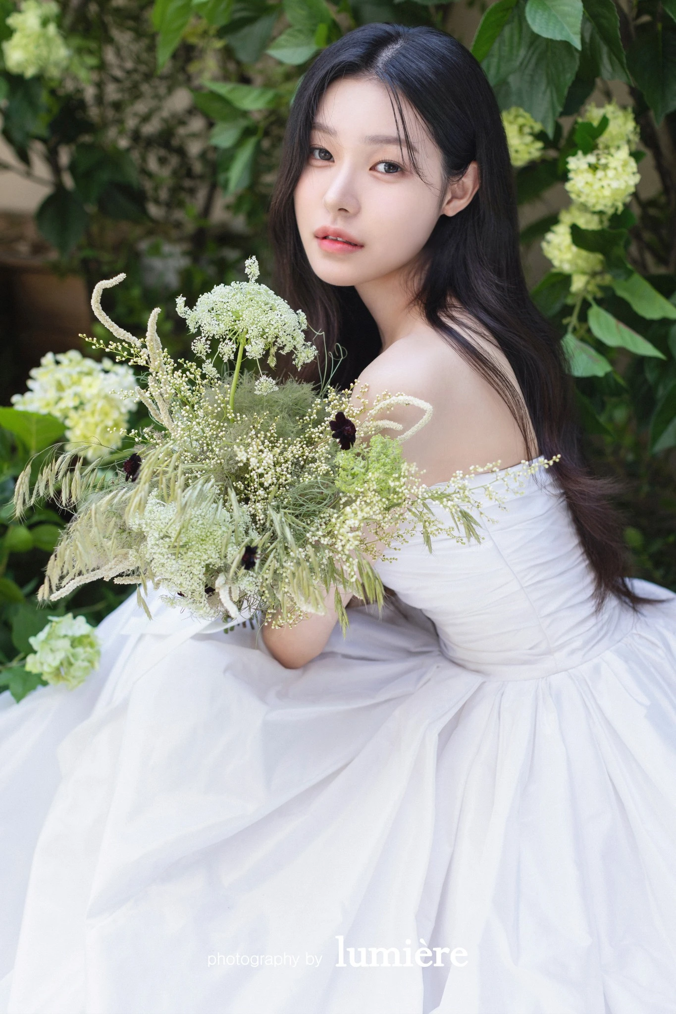 초록 잎이 짙은 꽃 배경에서 웨딩드레스를 입은 여성 단독이 부케를 들고 어깨 너머로 바라봄 모습이다. 그린 부케·흰 꽃이 함께 보인다.