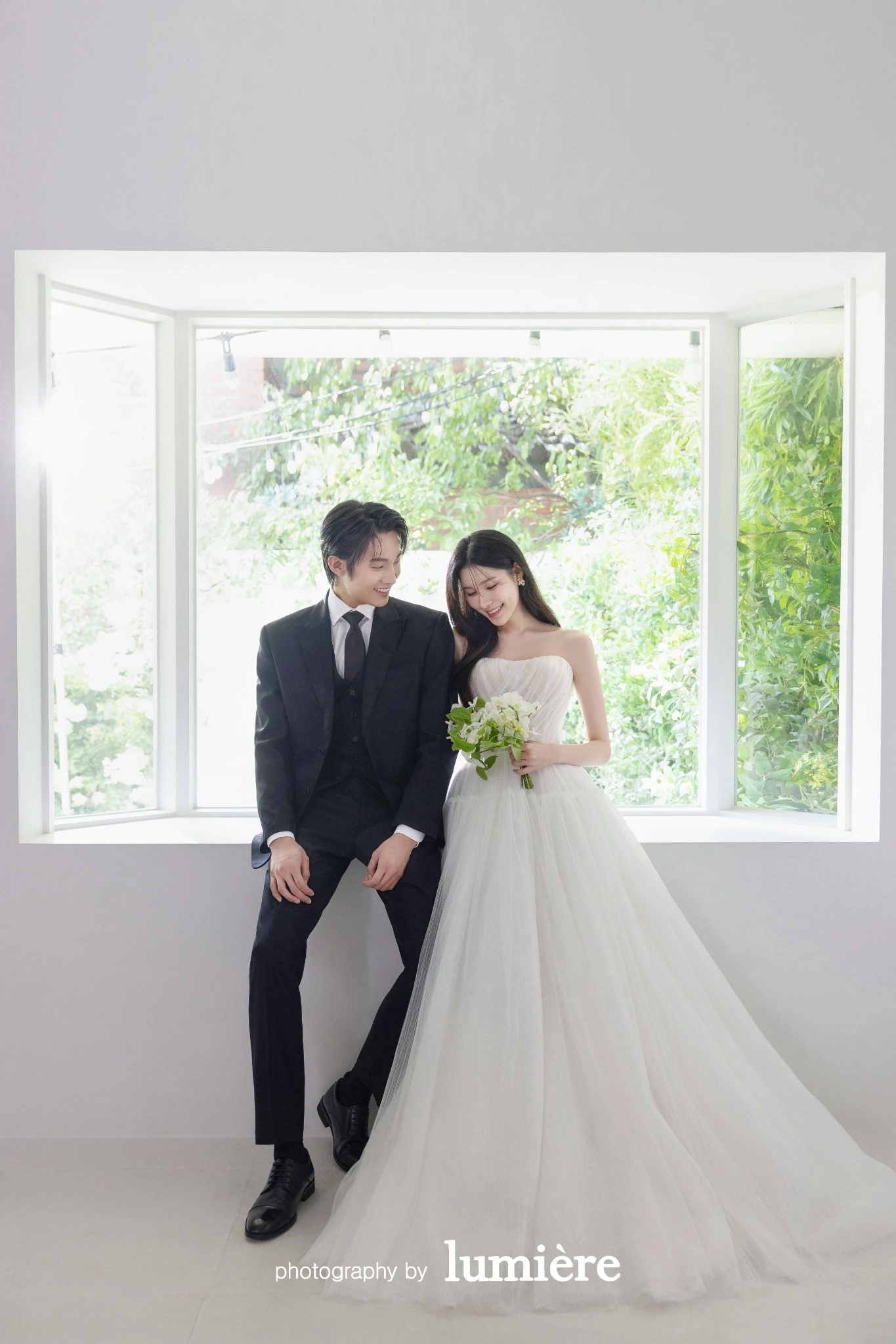 큰 창이 있는 흰 실내 공간에서 웨딩 의상의 남녀 모델이 창틀에 앉아 서로를 향함 모습이다. 부케·통창이 함께 보인다.