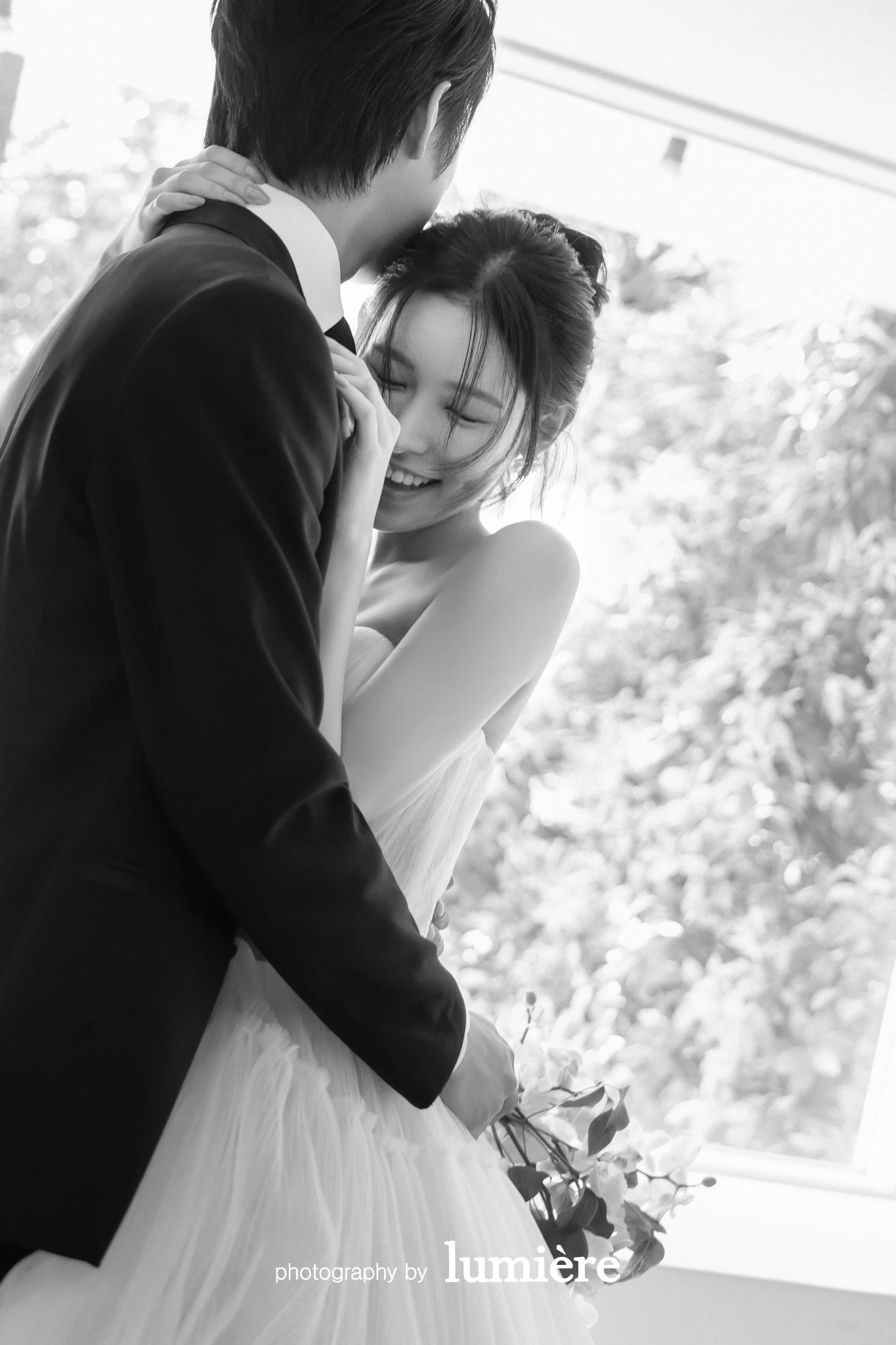 창가 근처 실내 공간에서 웨딩 의상의 남녀 모델이 남녀가 가까이 기대어 있음 모습이다. 부케·창문이 함께 보인다.