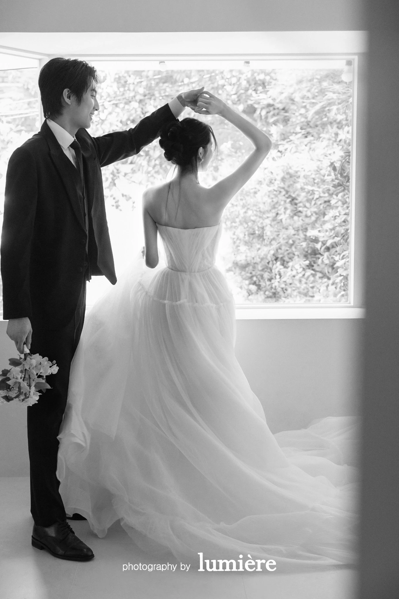 창가가 보이는 실내 공간에서 웨딩 의상의 남녀 모델이 남성이 여성의 손을 들어 회전 동작을 만듦 모습이다. 부케·창문이 함께 보인다.