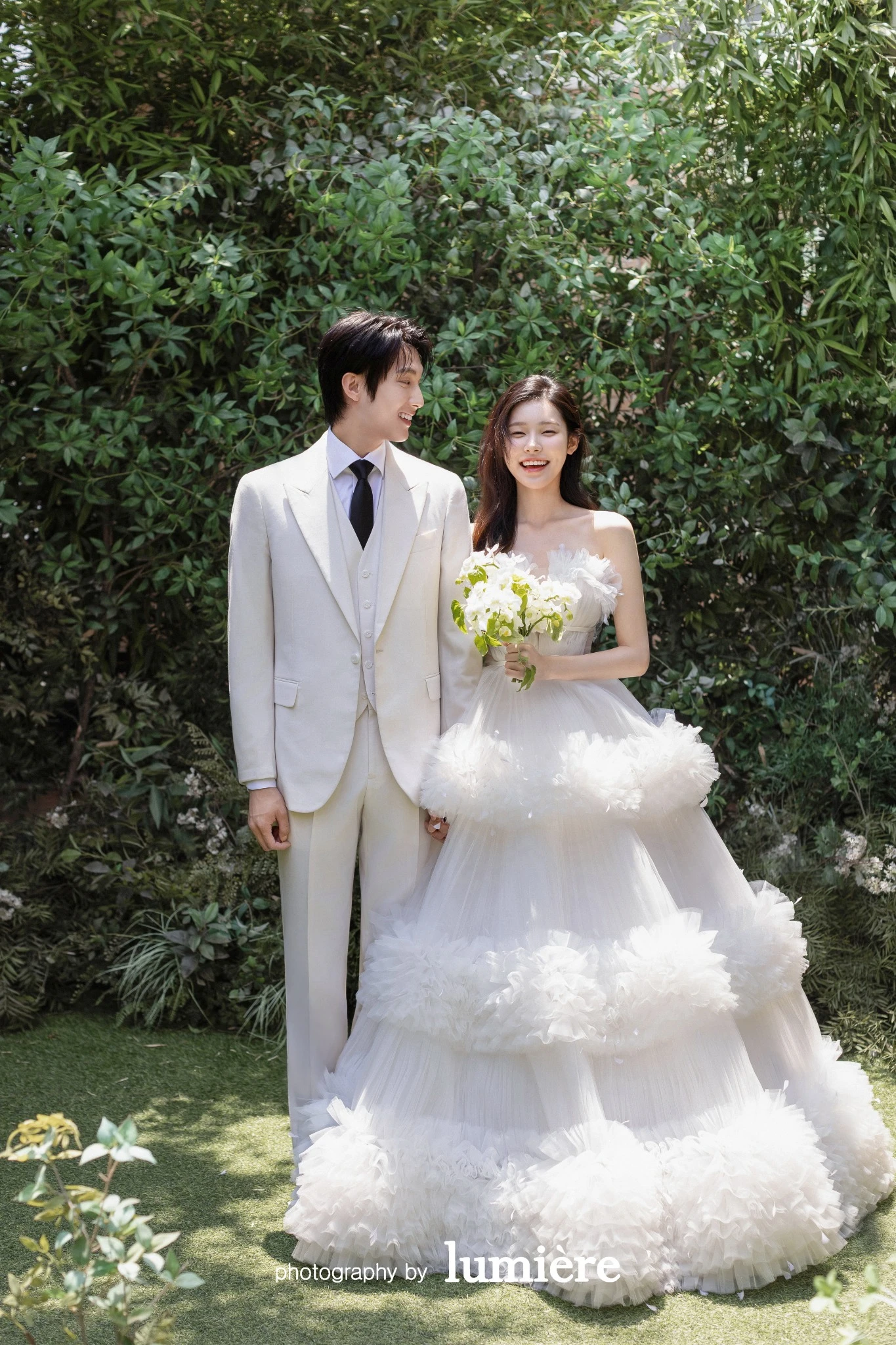 초록 잎이 많은 야외형 배경에서 웨딩 의상의 남녀 모델이 나란히 서서 카메라 방향을 바라봄 모습이다. 부케·화이트 예복이 함께 보인다.