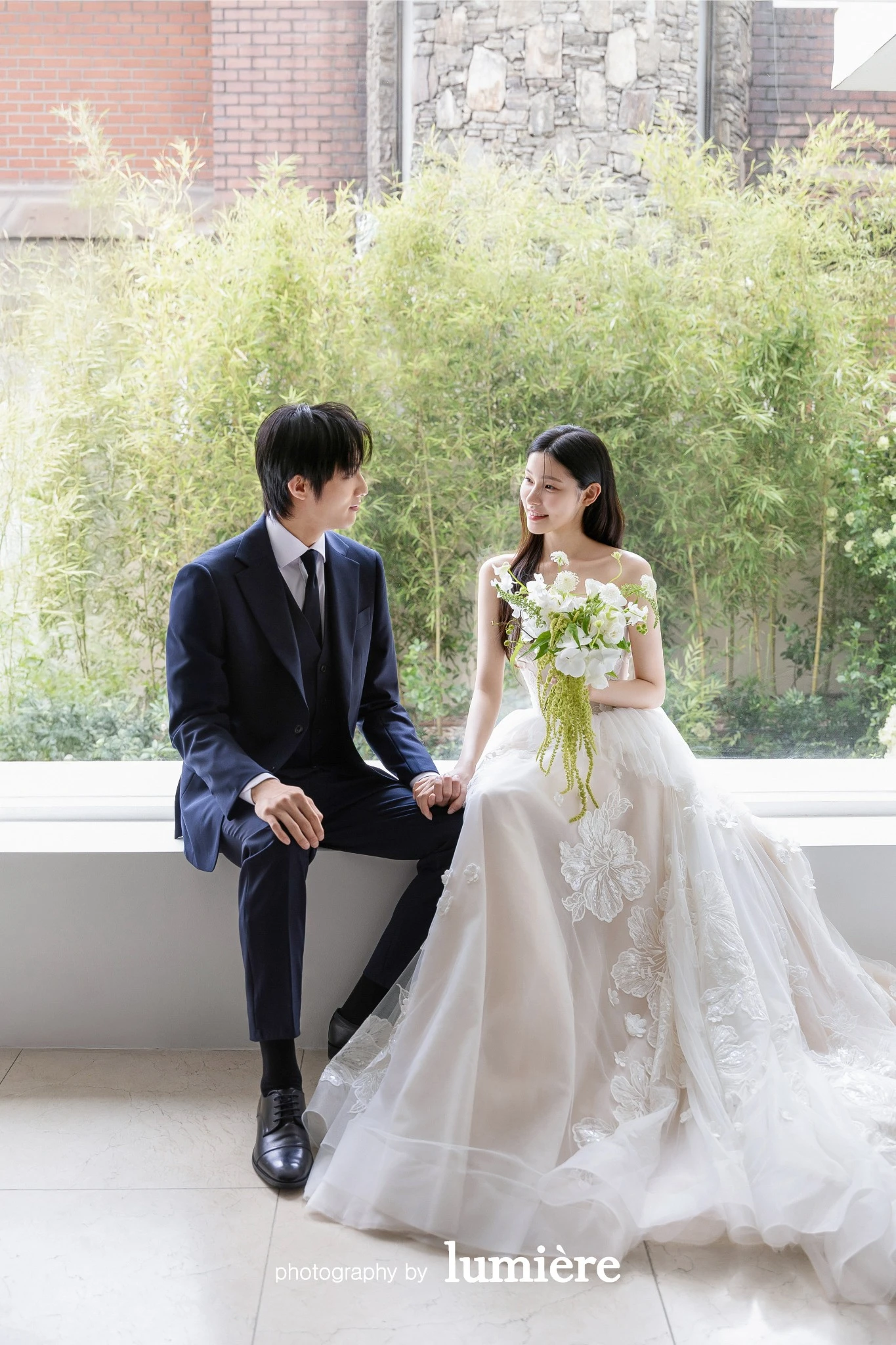 큰 창과 초록 배경이 보이는 실내에서 웨딩 의상의 남녀 모델이 창가에 앉아 손을 잡고 마주 봄 모습이다. 부케·창문이 함께 보인다.