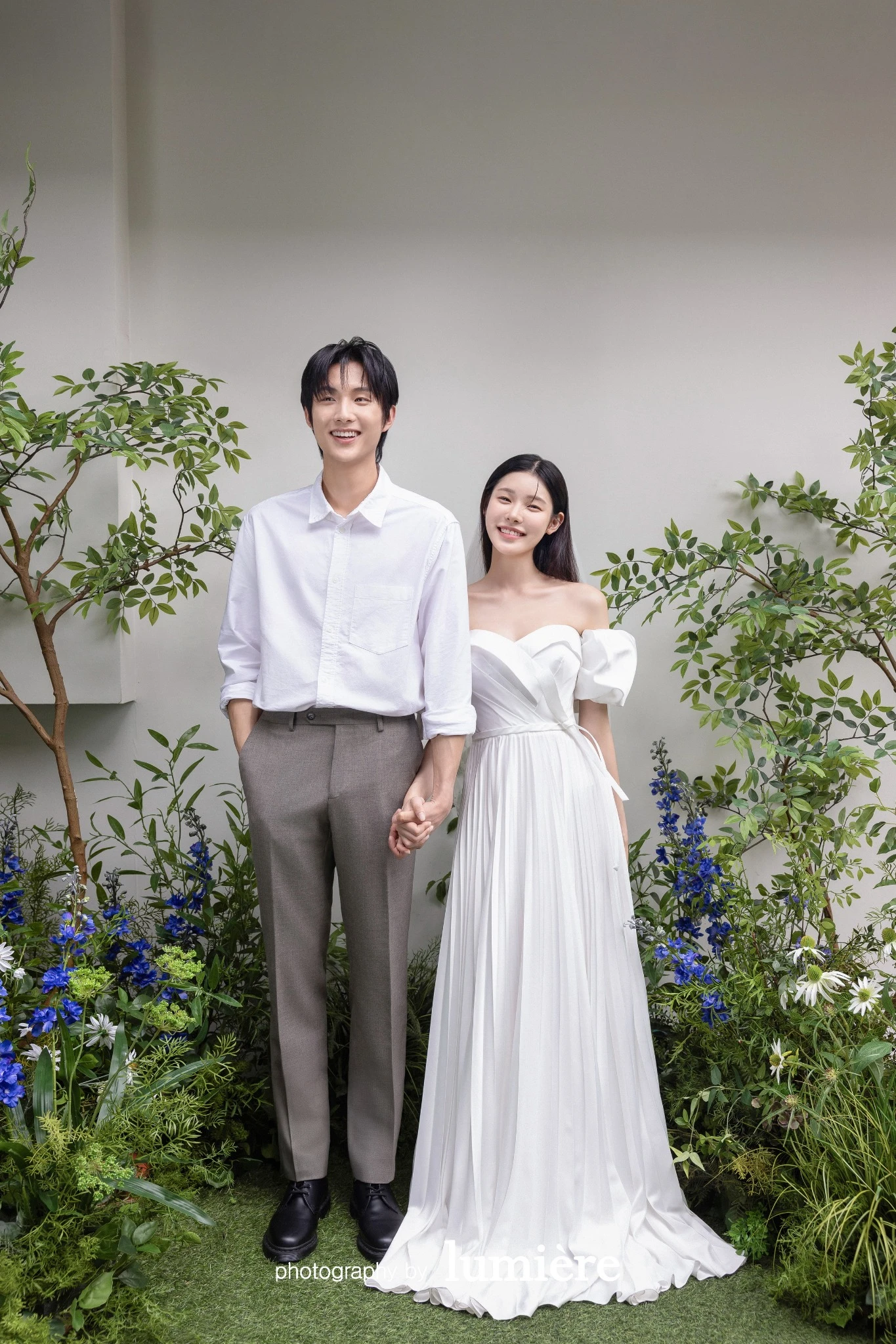 초록 식물이 있는 실내외 경계 공간에서 캐주얼 의상의 남녀 모델이 손을 잡고 서서 미소를 보임 모습이다. 흰 원피스·흰 셔츠가 함께 보인다.