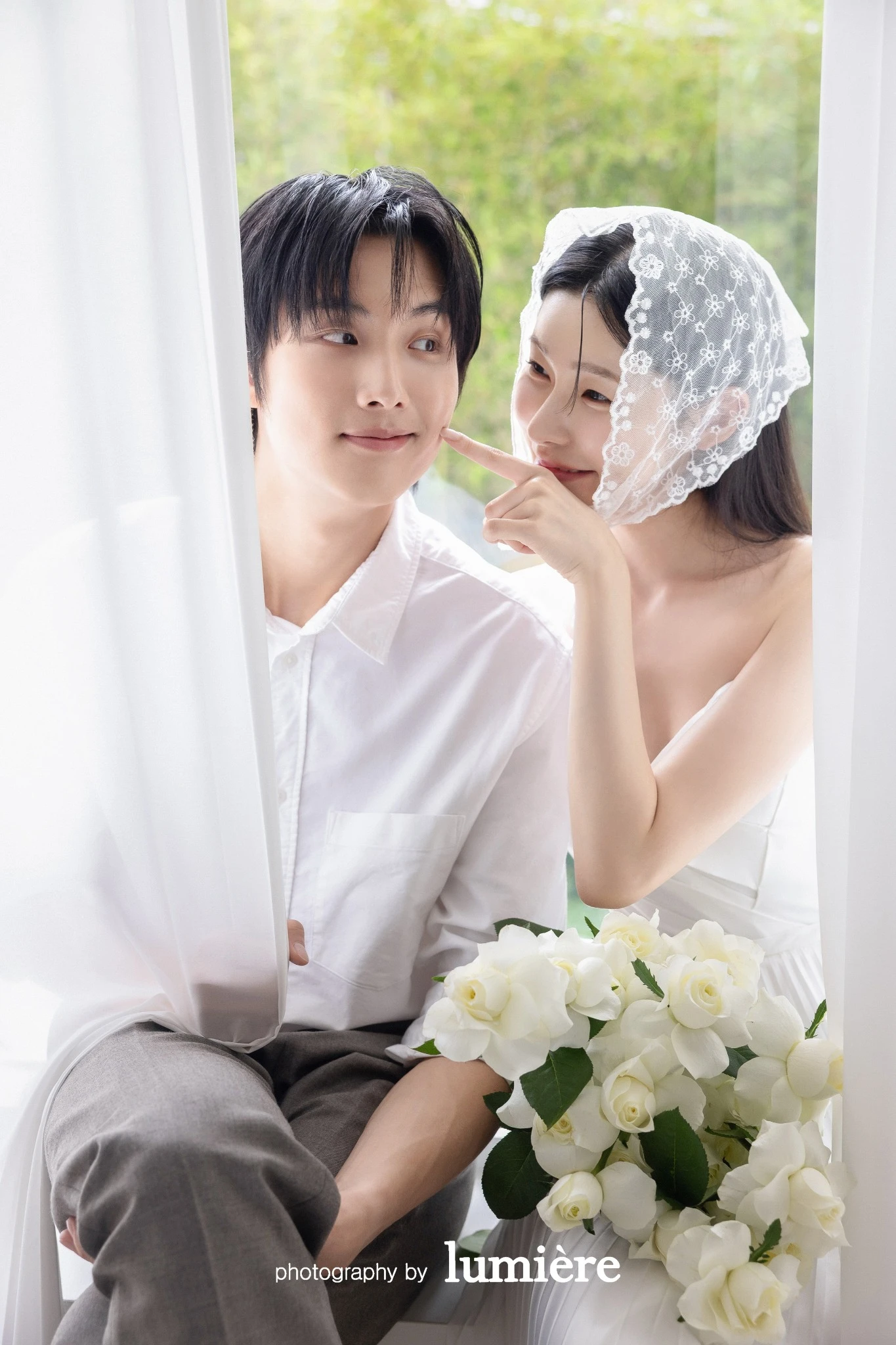 흰 커튼과 창가가 있는 밝은 공간에서 캐주얼 의상의 남녀 모델이 커튼 사이에서 서로 가까이 앉아 있음 모습이다. 흰 커튼·흰 장미 부케가 함께 보인다.