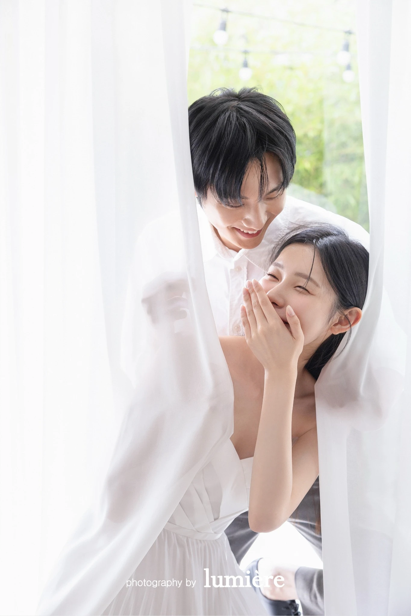 흰 커튼 사이의 밝은 창가에서 캐주얼 의상의 남녀 모델이 커튼 사이에서 웃는 표정을 보임 모습이다. 흰 커튼·화이트 의상이 함께 보인다.