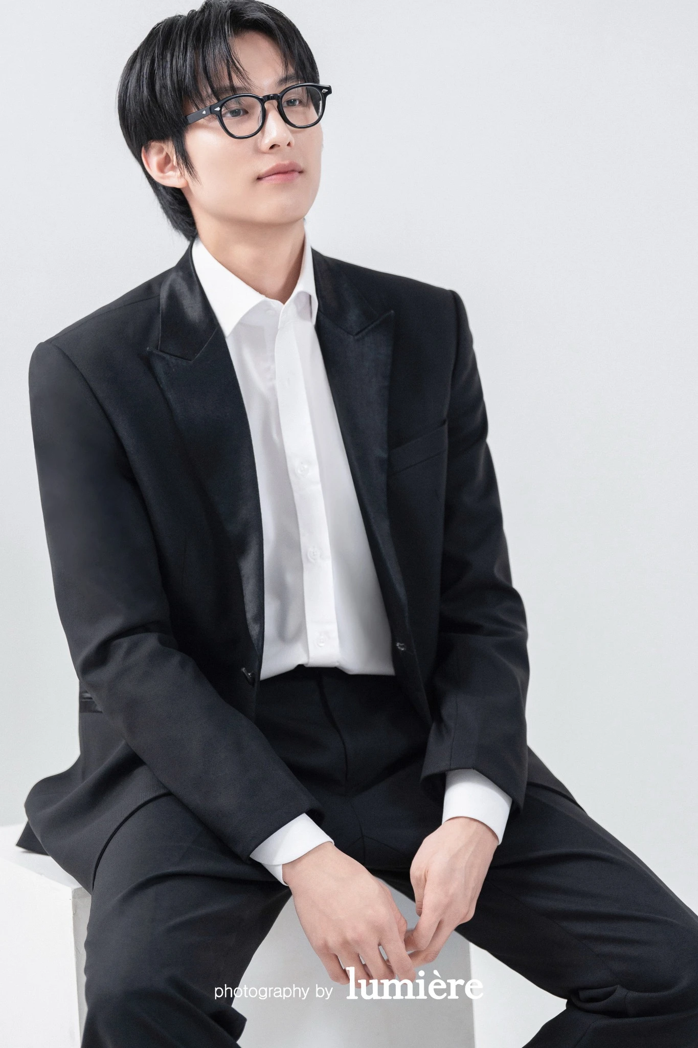 흰 배경의 미니멀 실내 세트에서 검정 예복을 입은 남성 단독이 안경을 쓰고 앉아 있음 모습이다. 안경·검정 예복이 함께 보인다.