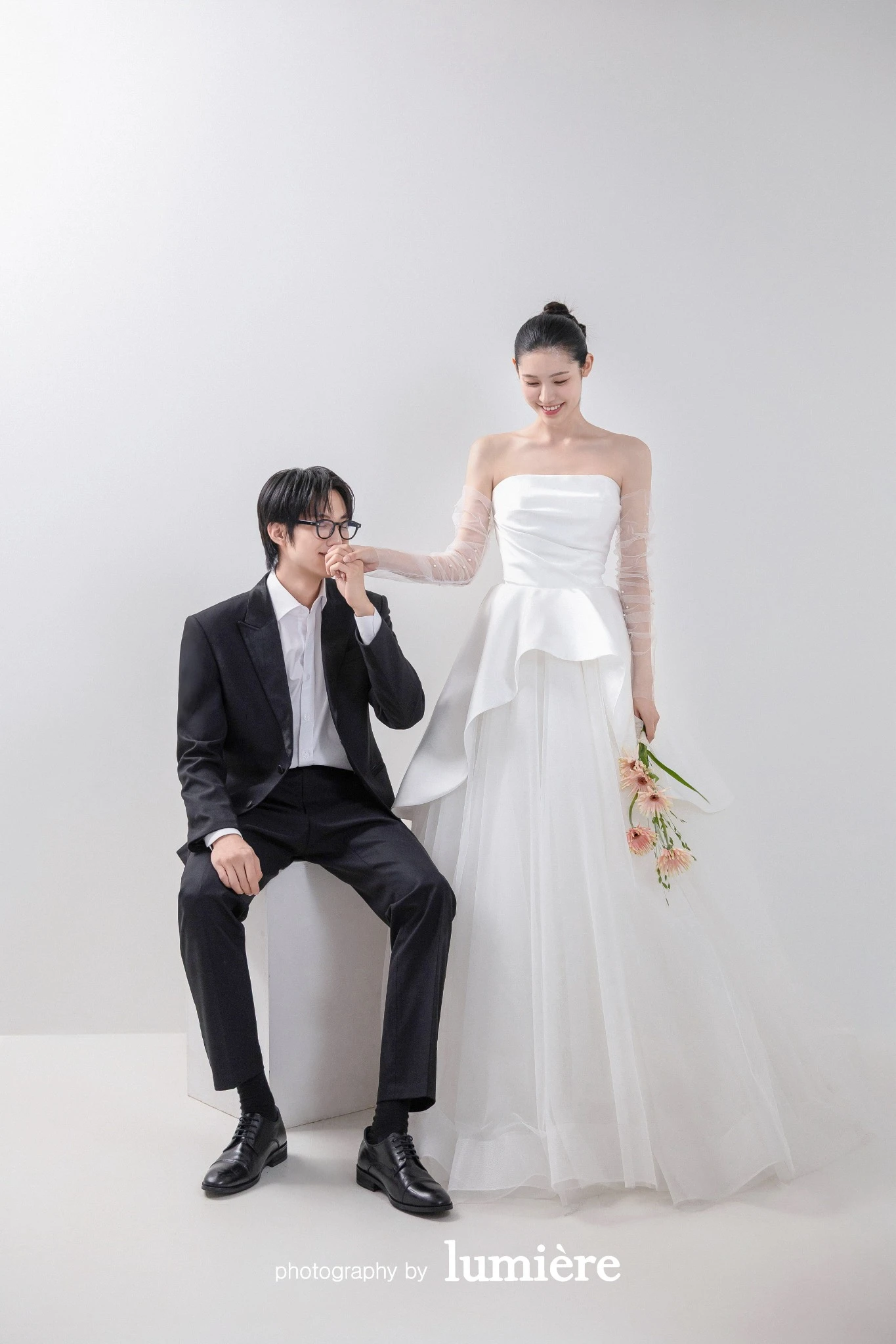 흰 배경의 미니멀 실내 세트에서 웨딩 의상의 남녀 모델이 남성이 여성의 손등에 입을 맞춤 모습이다. 화이트 드레스·검정 예복이 함께 보인다.