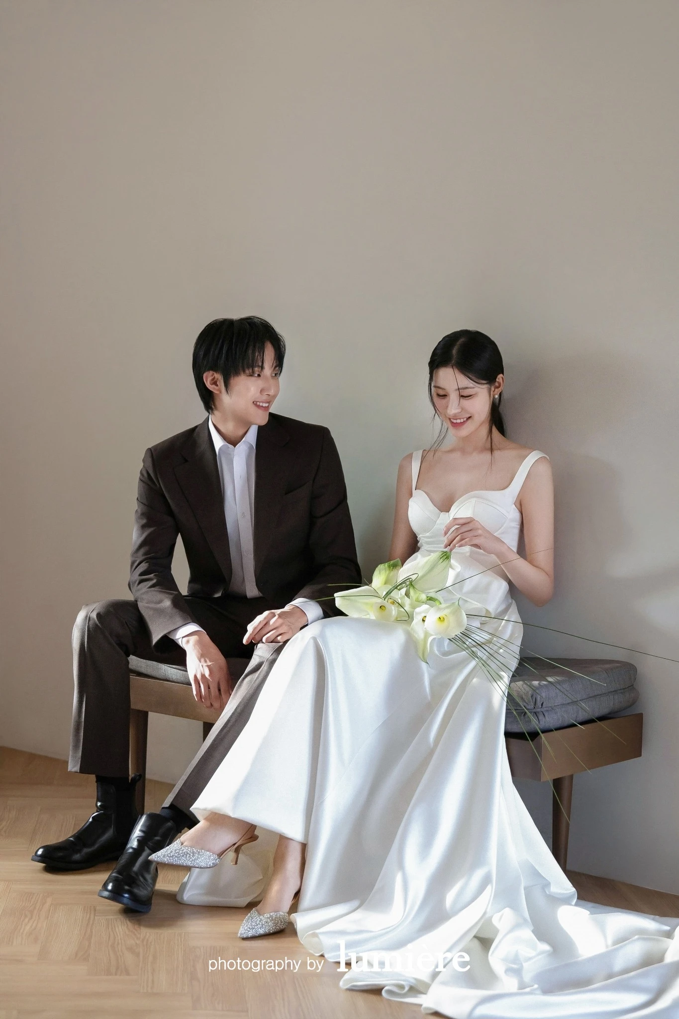 베이지 벽과 벤치가 있는 실내 세트에서 웨딩 의상의 남녀 모델이 벤치에 앉아 서로를 바라봄 모습이다. 칼라꽃 부케·벤치가 함께 보인다.