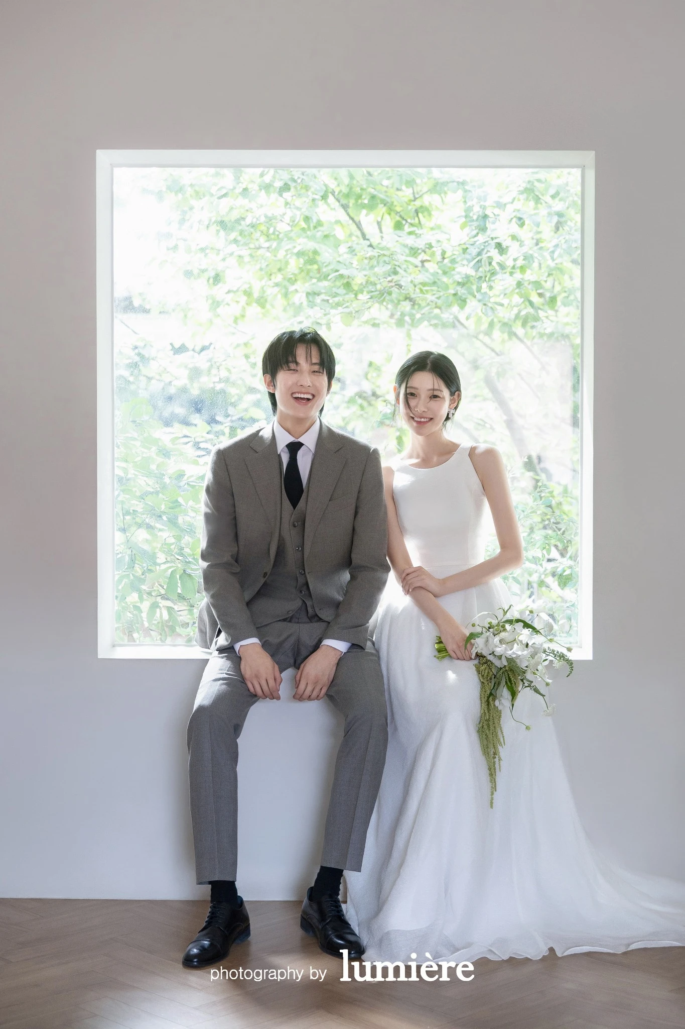 큰 창과 나무바닥이 있는 실내 공간에서 웨딩 의상의 남녀 모델이 창틀에 앉아 카메라 쪽을 향함 모습이다. 부케·창문이 함께 보인다.
