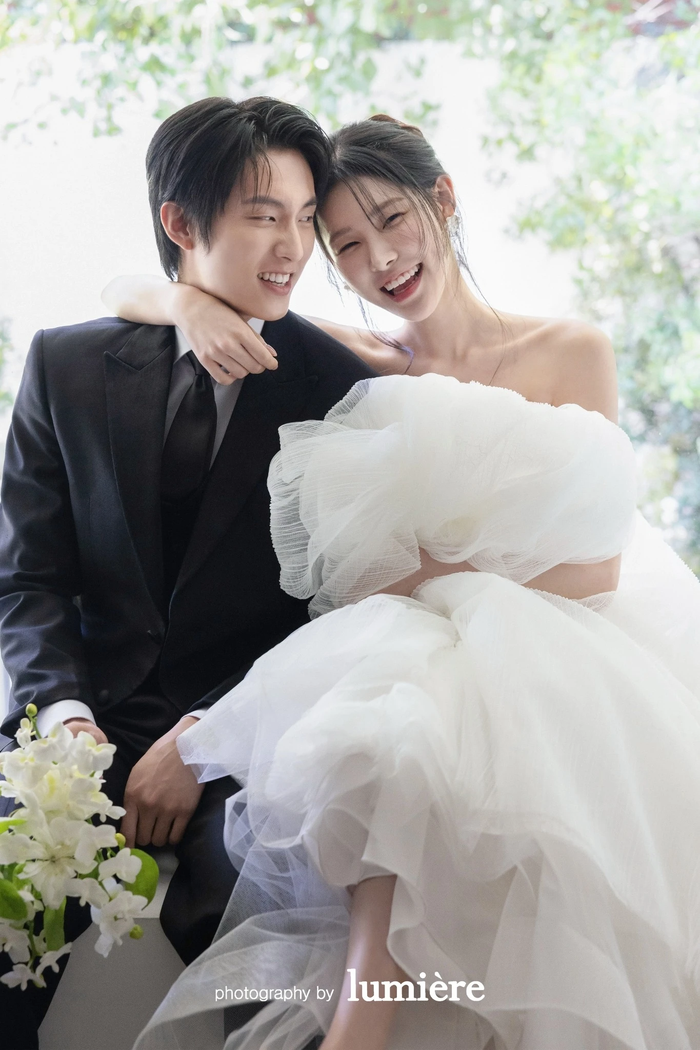 밝은 창가 실내 공간에서 웨딩 의상의 남녀 모델이 가까이 앉아 웃는 표정을 보임 모습이다. 부케·화이트 드레스가 함께 보인다.