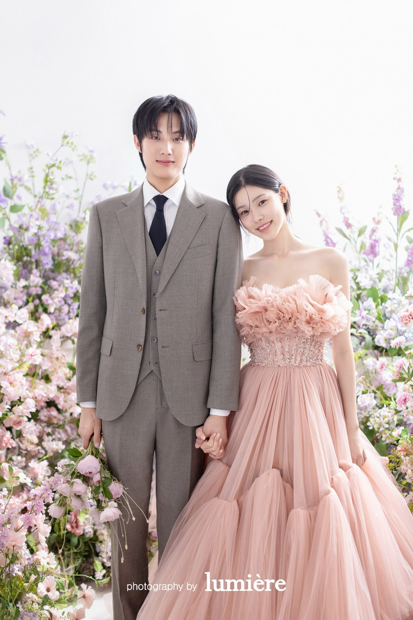 분홍 꽃이 있는 밝은 실내 꽃 세트에서 웨딩 의상의 남녀 모델이 나란히 서서 손을 잡음 모습이다. 분홍 드레스·회색 예복이 함께 보인다.