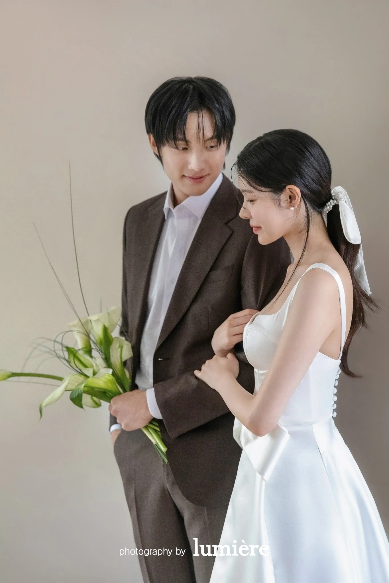 베이지 벽이 있는 실내 세트에서 웨딩 의상의 남녀 모델이 나란히 서서 서로를 바라봄 모습이다. 칼라꽃·브라운 예복이 함께 보인다.