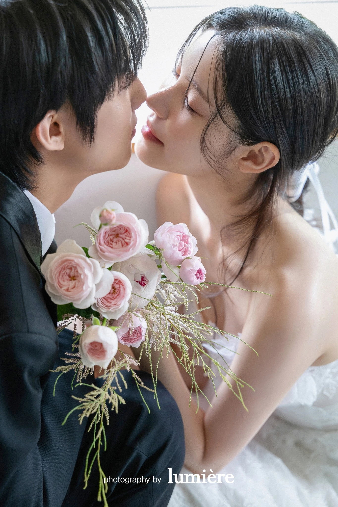 밝은 창가 또는 실내 공간에서 웨딩 의상의 남녀 모델이 부케를 사이에 두고 가까이 마주함 모습이다. 분홍 장미 부케·예복이 함께 보인다.