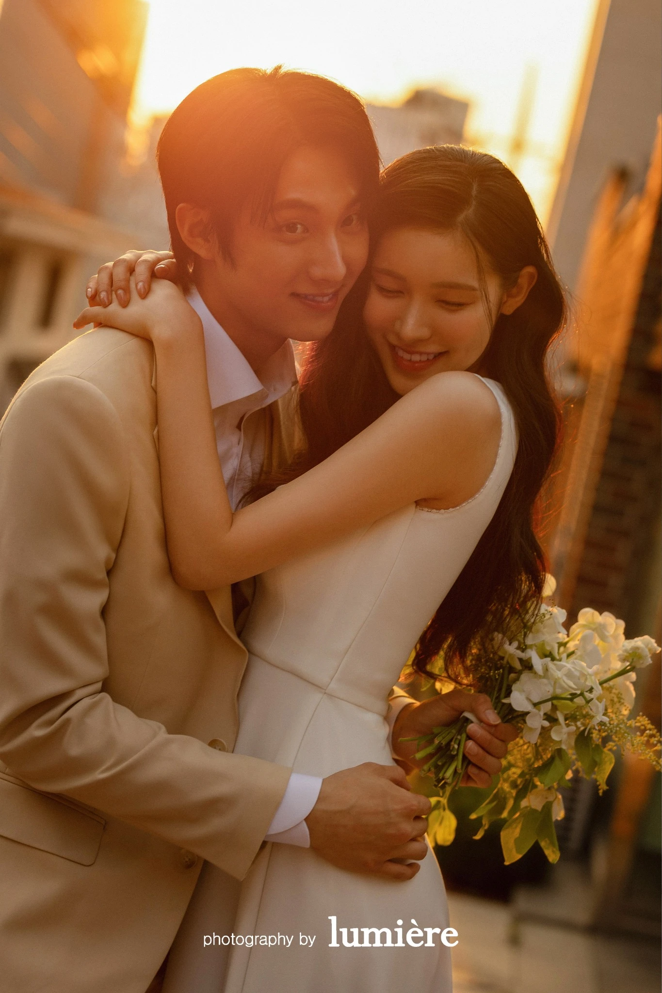 석양빛이 들어오는 도로형 야외 공간에서 웨딩 의상의 남녀 모델이 가까이 안고 카메라 방향을 함께 바라봄 모습이다. 부케·베이지 예복이 함께 보인다.