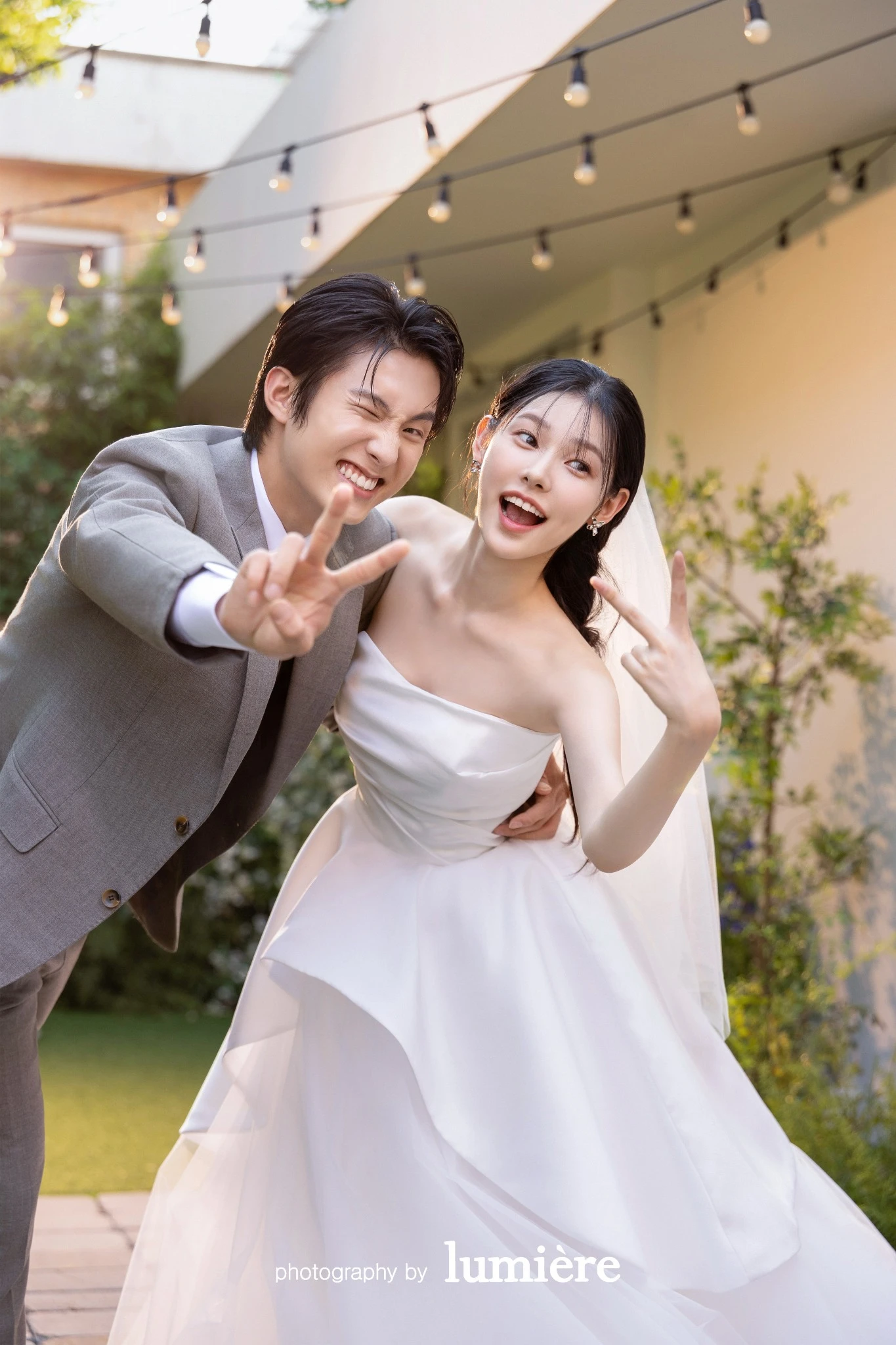 야외 정원형 공간에서 웨딩 의상의 남녀 모델이 카메라 쪽으로 다가와 손가락 브이 포즈를 보임 모습이다. 베일·회색 예복이 함께 보인다.