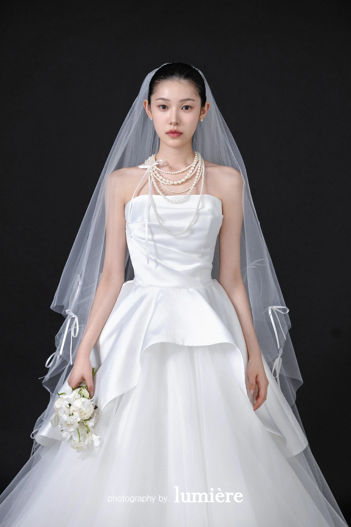 검은 배경의 스튜디오 세트에서 베일을 착용한 웨딩 의상의 남녀 모델이 손을 잡고 나란히 서 있음 모습이다. 베일·흰 부케가 함께 보인다.