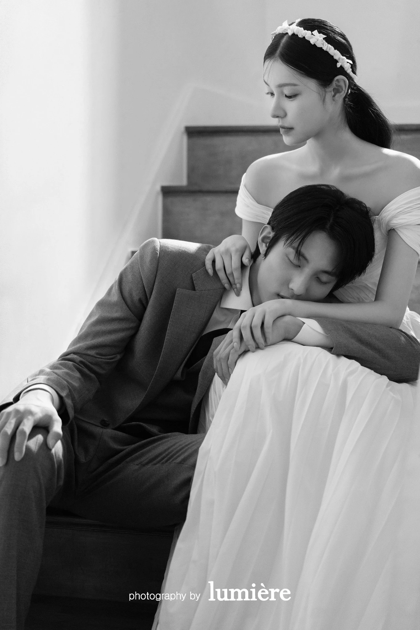 계단 옆 실내 공간에서 웨딩 의상의 남녀 모델이 남성이 여성의 무릎 근처에 기대어 있음 모습이다. 계단·드레스가 함께 보인다.
