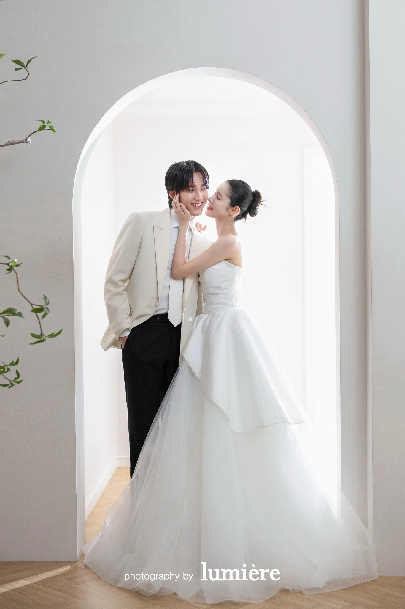 아치형 창문이 있는 밝은 실내 공간에서 웨딩 의상의 남녀 모델이 아치 안에서 가까이 서서 서로를 향함 모습이다. 아치형 창·화이트 예복이 함께 보인다.