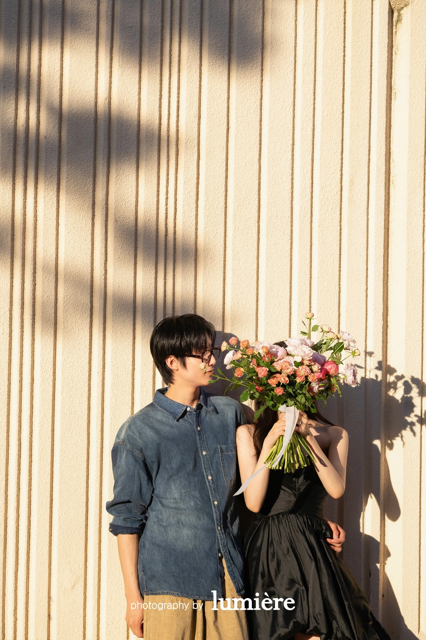 세로 홈이 있는 벽 앞 야외 공간에서 캐주얼 의상의 남녀 모델이 꽃다발로 얼굴 일부를 가리고 나란히 서 있음 모습이다. 꽃다발·데님 셔츠가 함께 보인다.