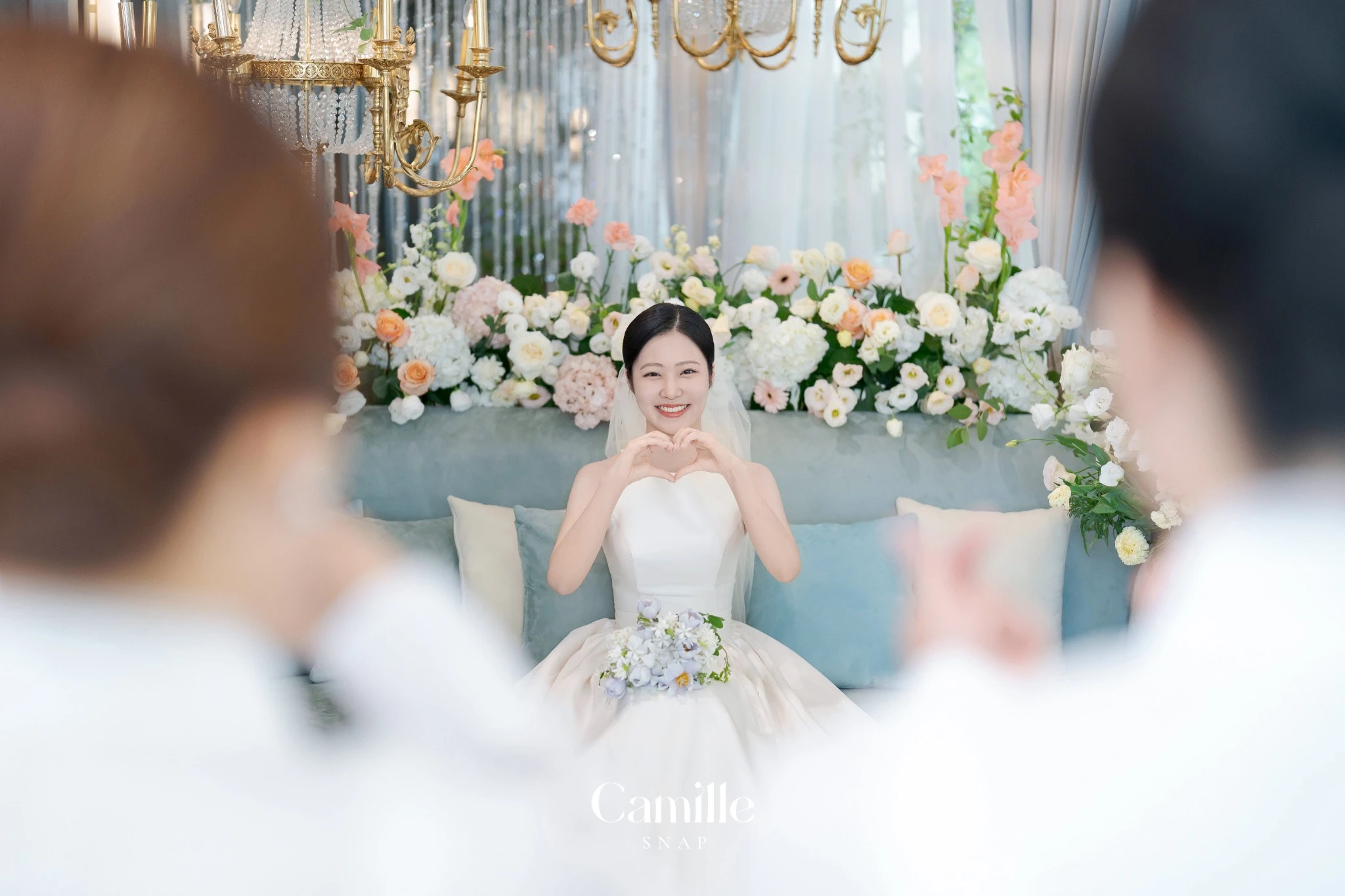 전경 인물 사이로 신부의 밝은 포즈가 보이는 자연스러운 컷입니다.