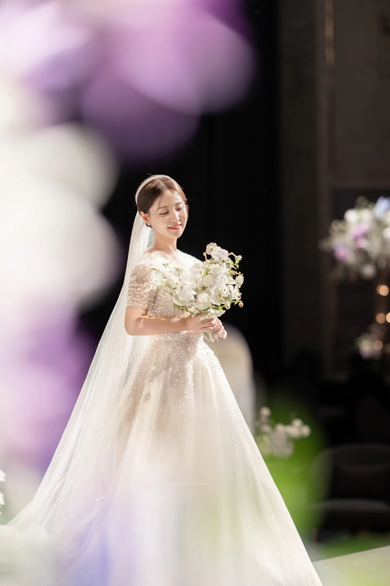 공간 중심 커플 컷으로, 높은 천장과 샹들리에가 있는 밝은 예식홀와 샹들리에과 기하학 천장 조명가 함께 보이는 장면입니다.