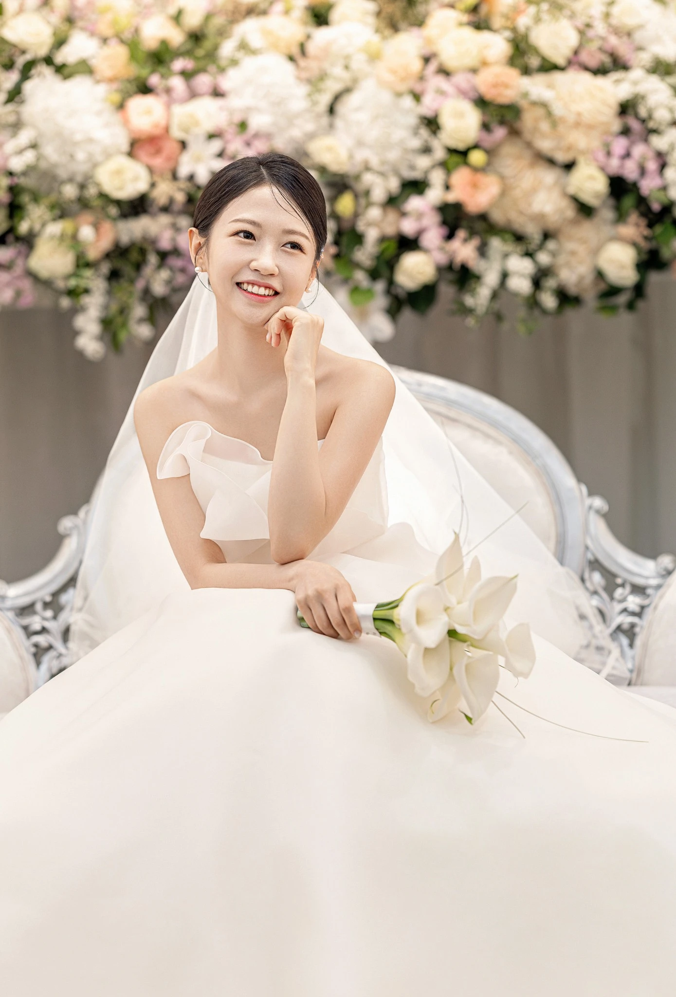 소파 커플 인물 컷으로, 화이트 소파와 파스텔 꽃벽 앞와 소파과 부케가 함께 보이는 장면입니다.