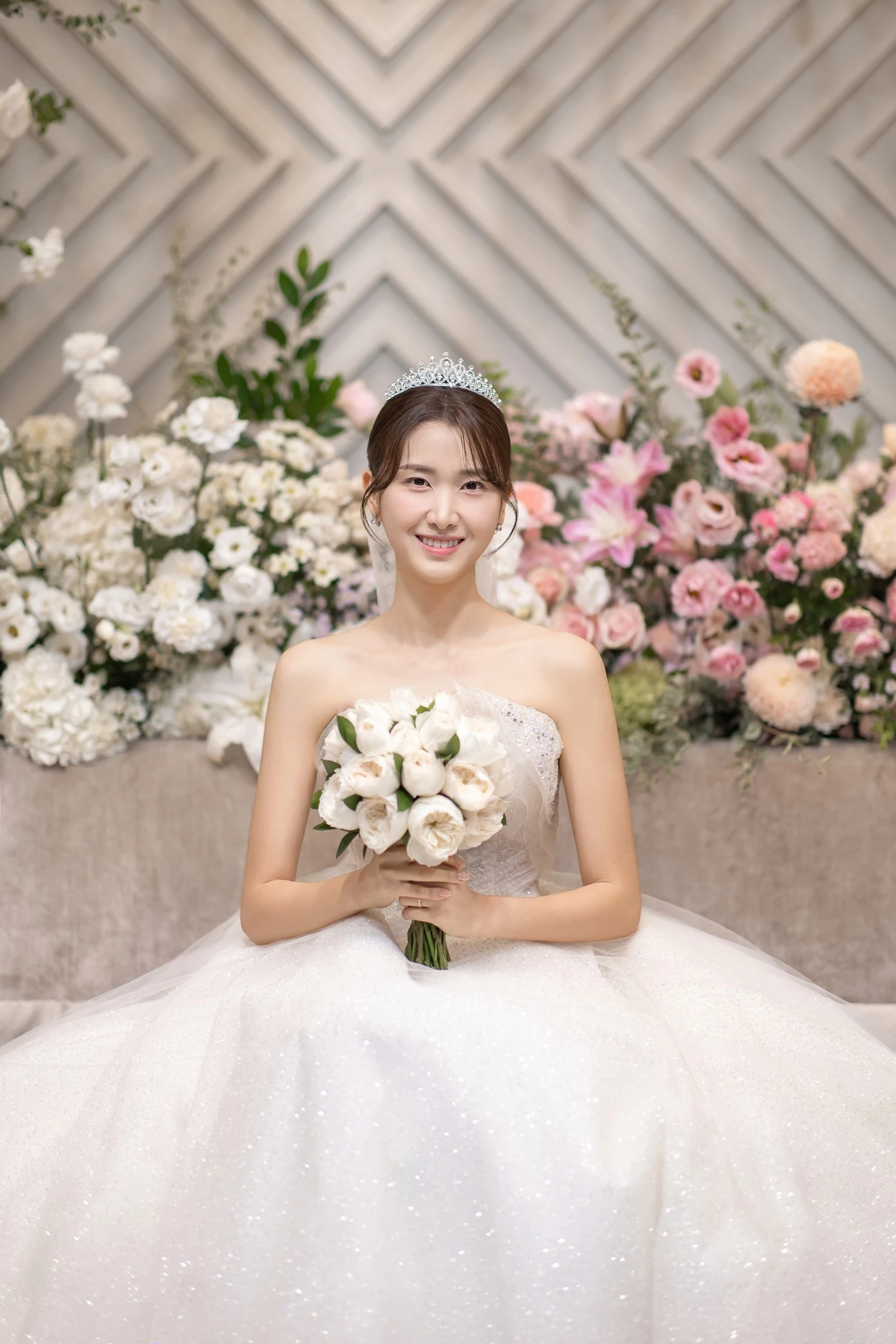 소파 커플 인물 컷으로, 꽃 장식이 많은 단상과 소파와 소파과 부케가 함께 보이는 장면입니다.