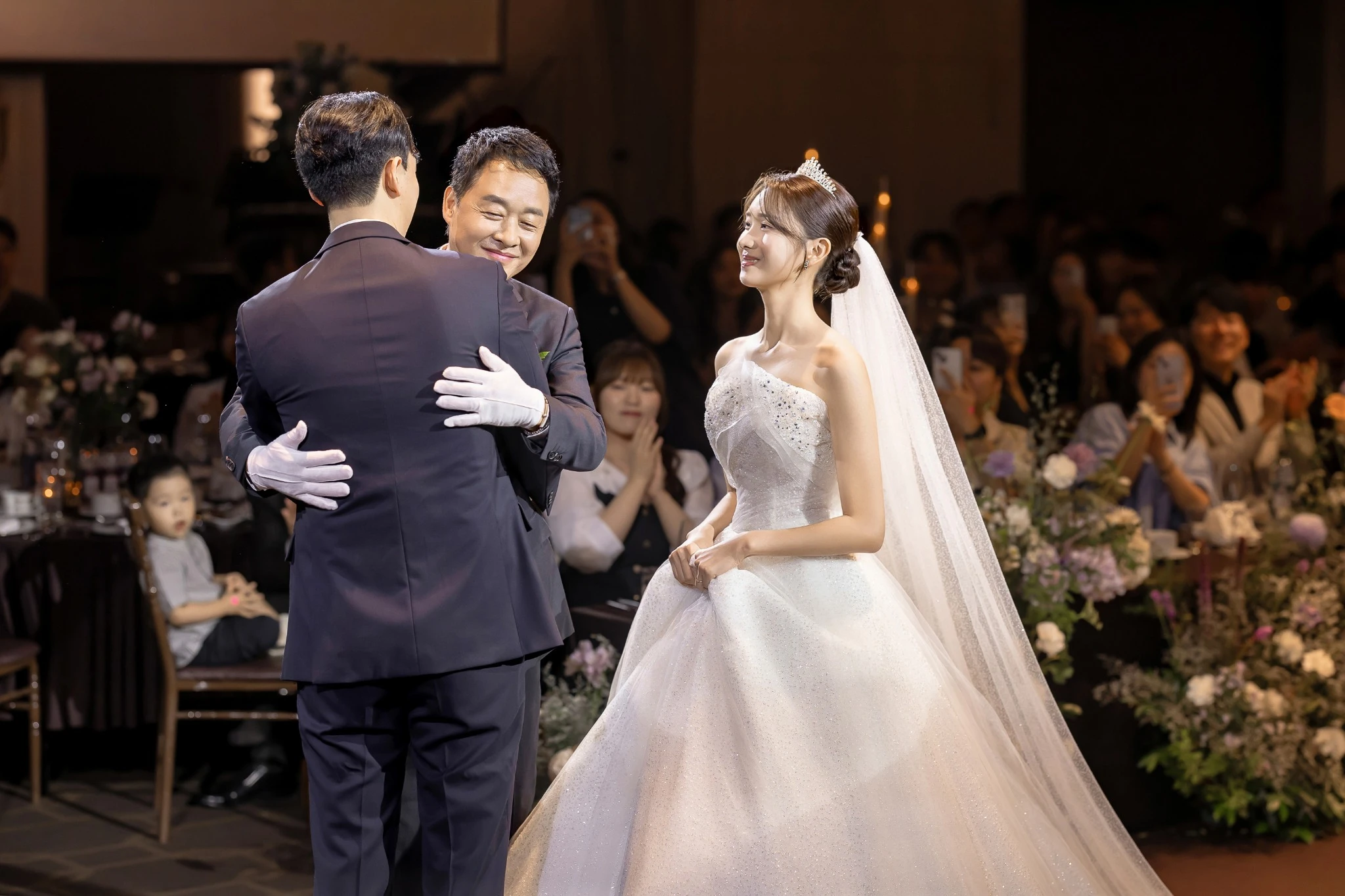 가족 동행 입장 장면으로, 하객이 둘러싼 어두운 버진로드와 하객와 촛불가 함께 보이는 장면입니다.