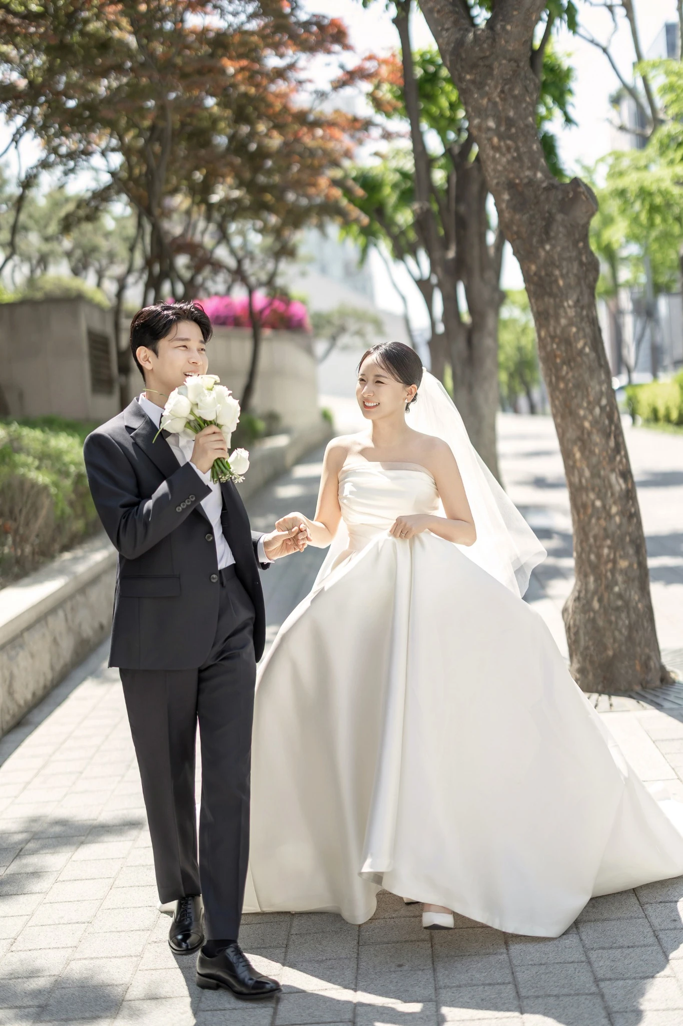 밝은 플라워 커플 컷으로, 밝은 꽃 장식과 흰 배경의 실내 공간와 부케과 긴 베일가 함께 보이는 장면입니다.