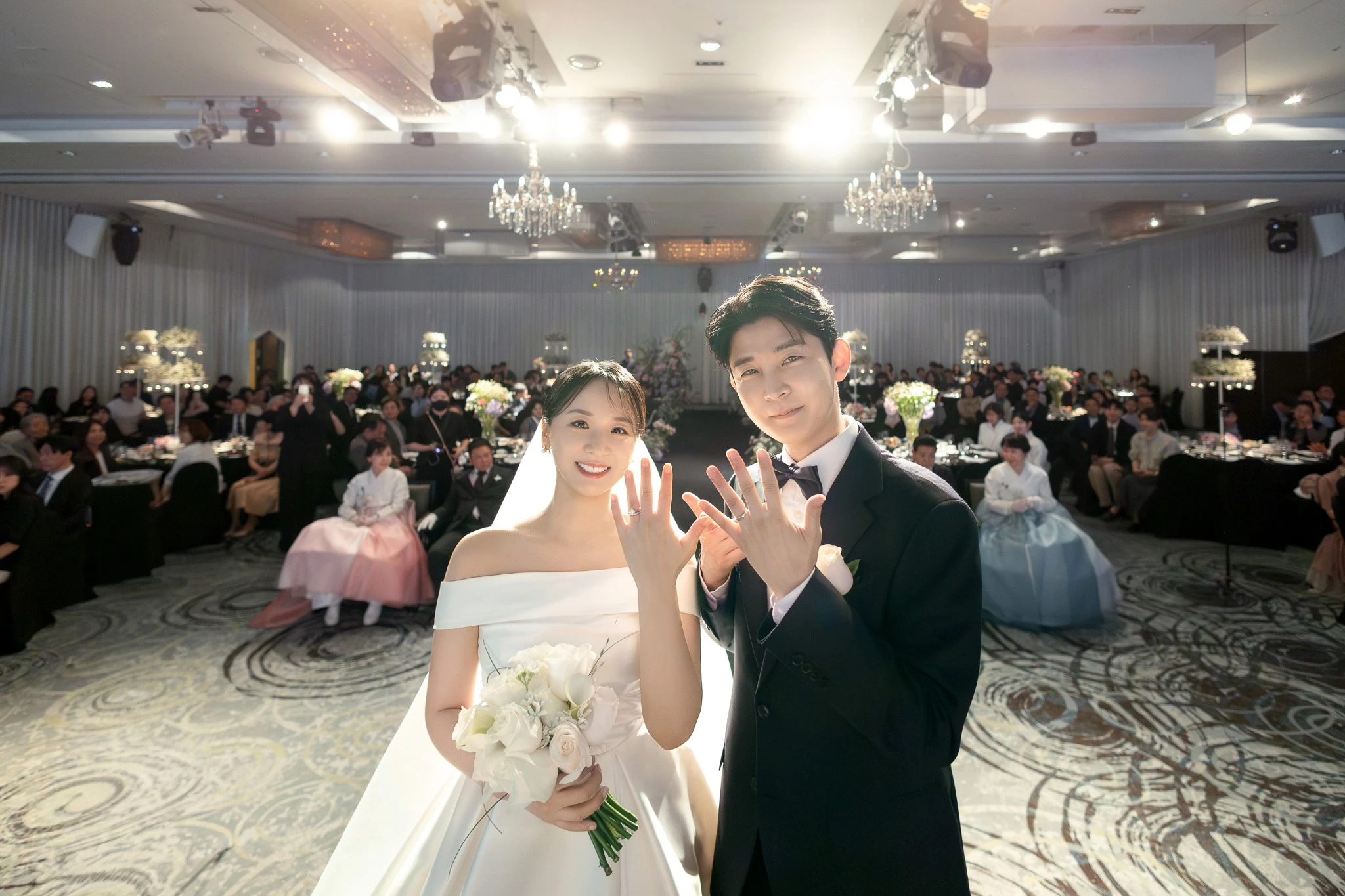 연회 토스트 인물 컷으로, 넓은 연회홀 중앙와 샴페인잔와 하객석가 함께 보이는 장면입니다.