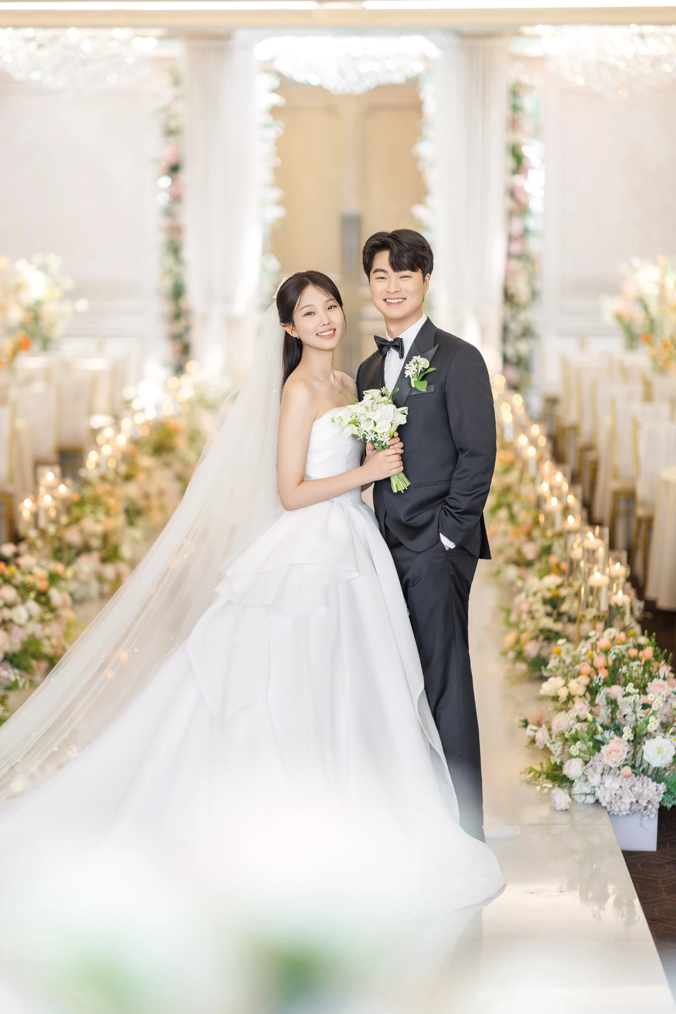 플라워 허그 커플 컷으로, 흰 꽃과 초록 잎이 많은 밝은 실내와 부케과 베일가 함께 보이는 장면입니다.