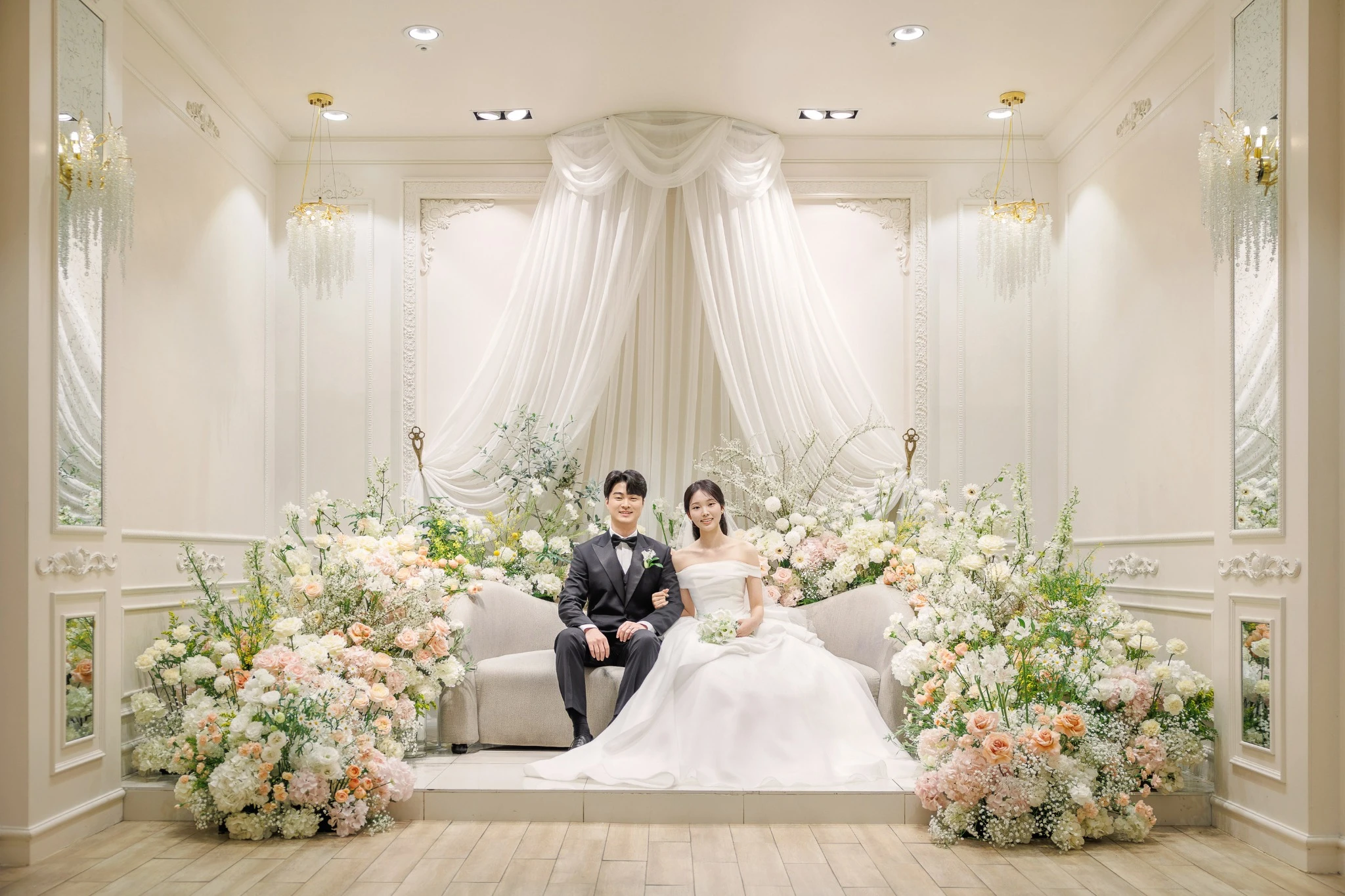 크림톤 단상 커플 컷으로, 크림색 벽과 드레이프 배경의 실내 단상와 드레이프 장식와 소파가 함께 보이는 장면입니다.