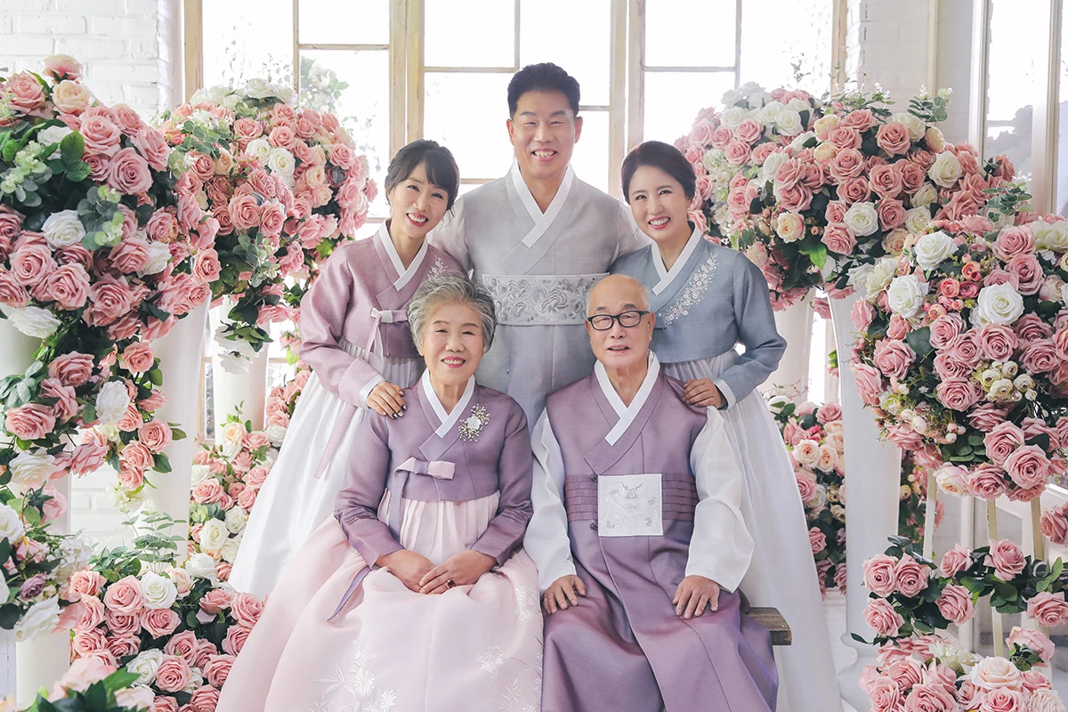 밝은 창과 흰 벽돌이 보이는 꽃 장식 실내에서 한복을 입은 성인 5명이 분홍색과 흰색 꽃 장식 사이에 앉고 선 사진이며, 풍성한 꽃 장식, 벤치가 장면의 구성을 보완합니다.