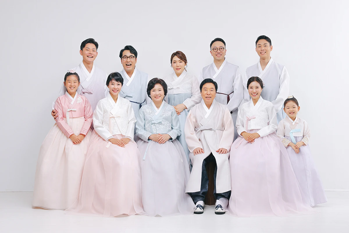 밝은 흰색 스튜디오 배경에서 한복을 입은 성인과 아동들이 흰 배경 앞에서 앉고 선 단체 사진이며, 낮은 의자와 흰 바닥가 장면의 구성을 보완합니다.