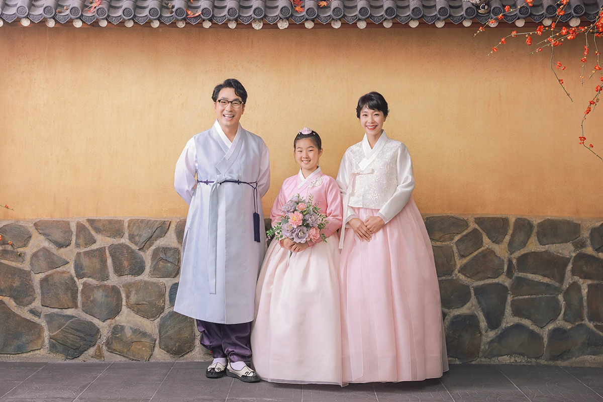 전통 담장과 기와 느낌의 배경에서 한복을 입은 성인 2명과 아동 1명이 담장 형태 배경 앞에 선 사진이며, 꽃다발, 돌담 장식가 장면의 구성을 보완합니다.