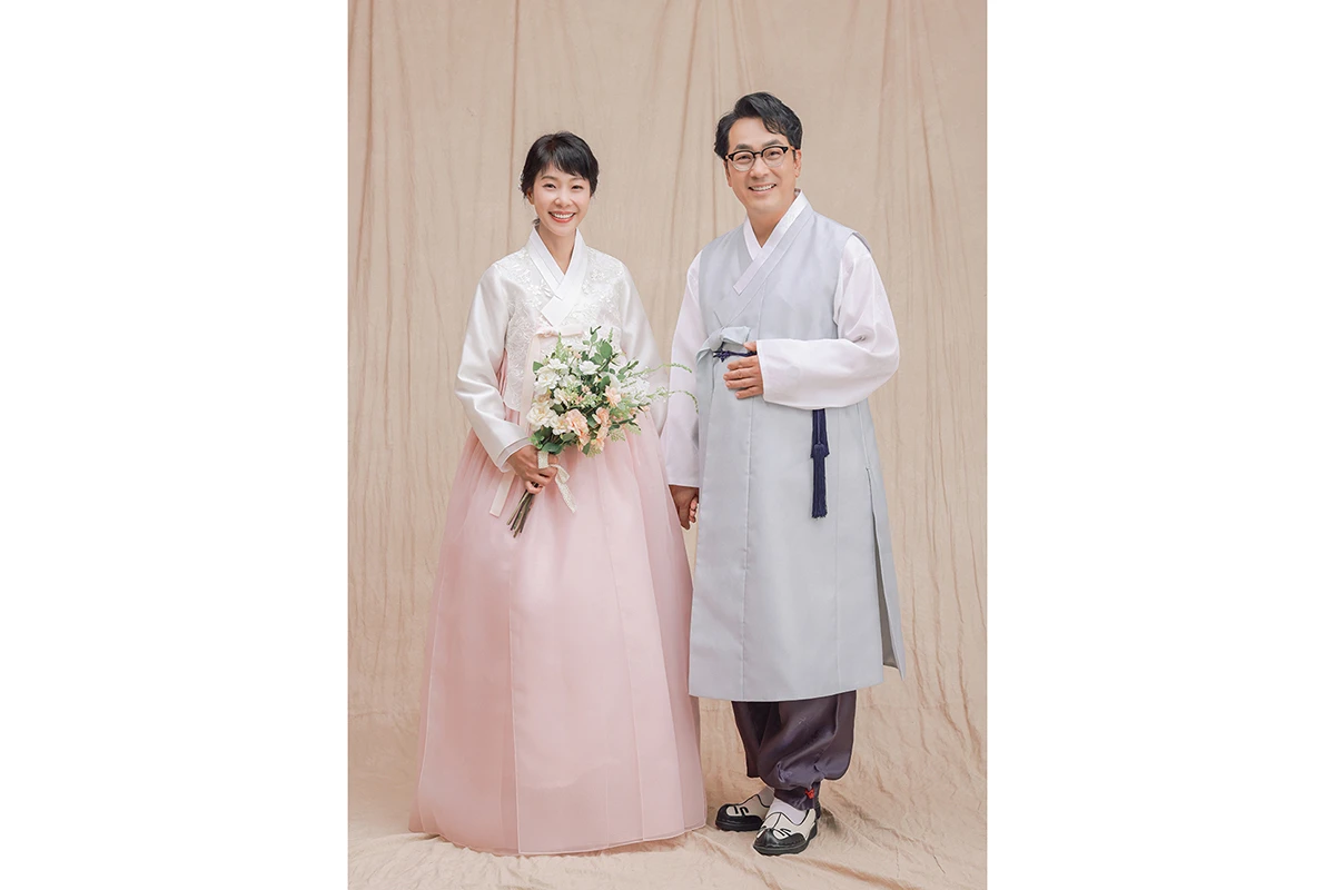 베이지 천 배경의 실내 스튜디오에서 한복을 입은 성인 2명이 베이지 천 배경 앞에서 손을 잡고 선 사진이며, 꽃다발과 천 배경가 장면의 구성을 보완합니다.