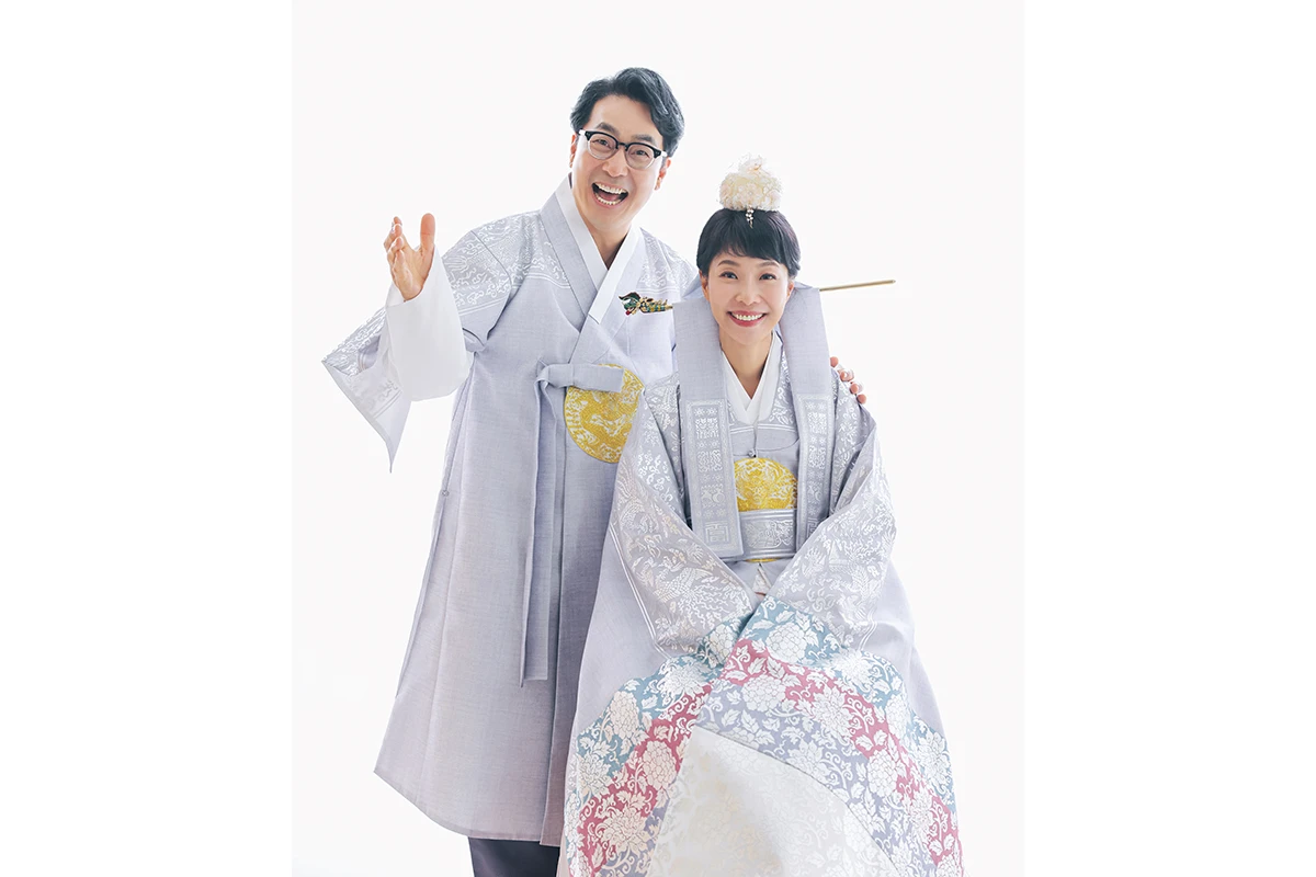 흰 배경의 밝은 스튜디오에서 한복을 입은 성인 2명이 흰 배경 앞에서 상반신 가까이 모인 사진이며, 한복 머리 장식과 넓은 소매가 장면의 구성을 보완합니다.