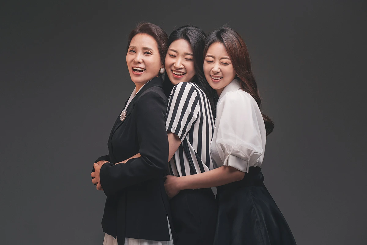 어두운 회색 배경의 스튜디오에서 검정과 흰색 의상의 성인 5명이 회색 배경 앞 테이블에 가까이 모인 사진이며, 흰색 테이블과 밀착 포즈가 장면의 구성을 보완합니다.