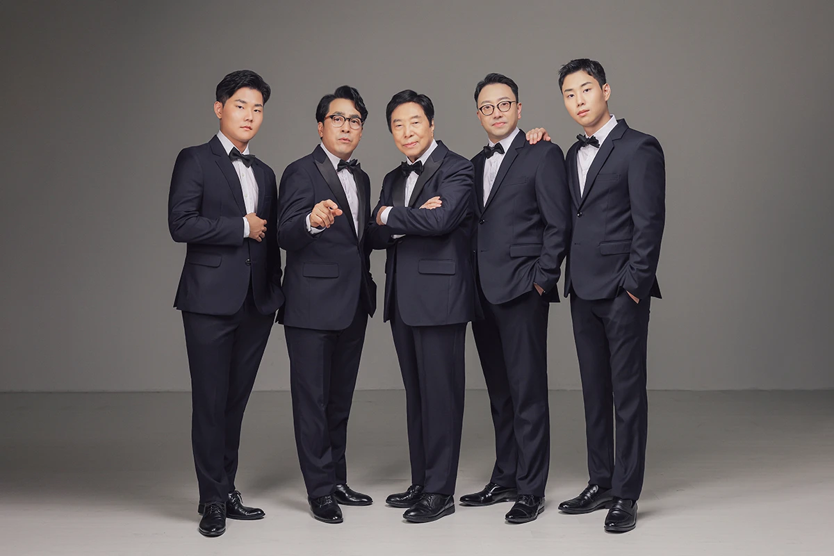 어두운 회색 스튜디오 배경에서 검정 의상을 입은 여성 3명이 어두운 회색 배경 앞에 가까이 선 사진이며, 의자나 소품 없이 인물 중심가 장면의 구성을 보완합니다.