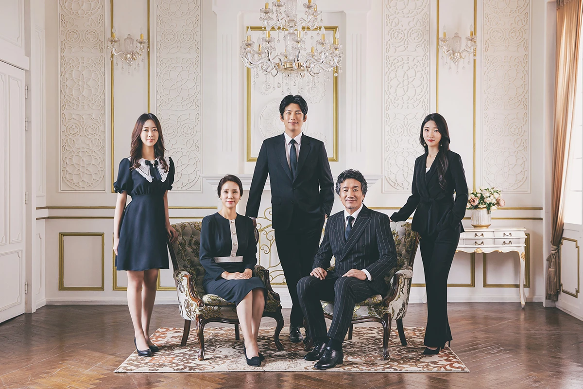 밝은 흰색 스튜디오 배경에서 검정과 흰색 의상의 여러 세대 인물과 아동, 반려견 두 마리가 흰 배경 앞에 모인 사진이며, 의자, 반려견, 흰 바닥가 장면의 구성을 보완합니다.