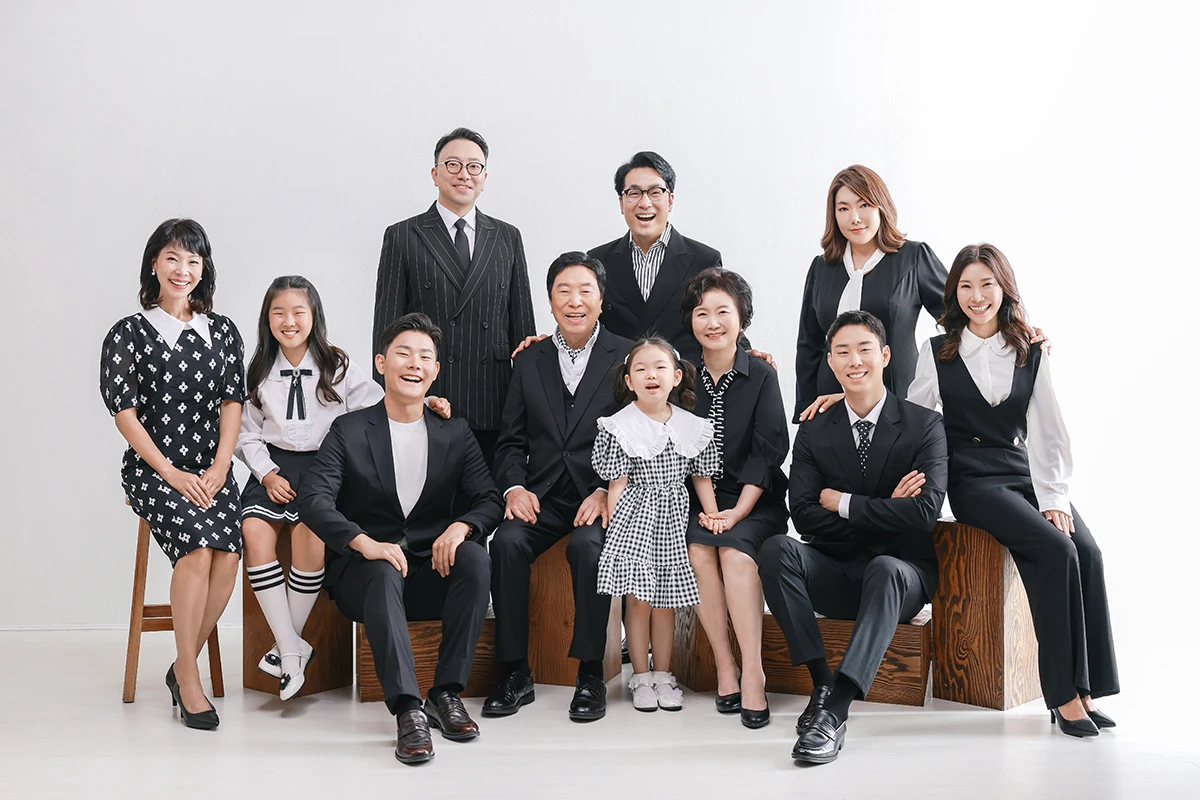 흰 벽돌과 큰 창이 있는 밝은 실내에서 검정과 흰색 의상을 입은 성인 3명과 아동 1명이 흰 벽돌 배경 앞에서 마주 보는 사진이며, 흰 의자와 창가 장면의 구성을 보완합니다.