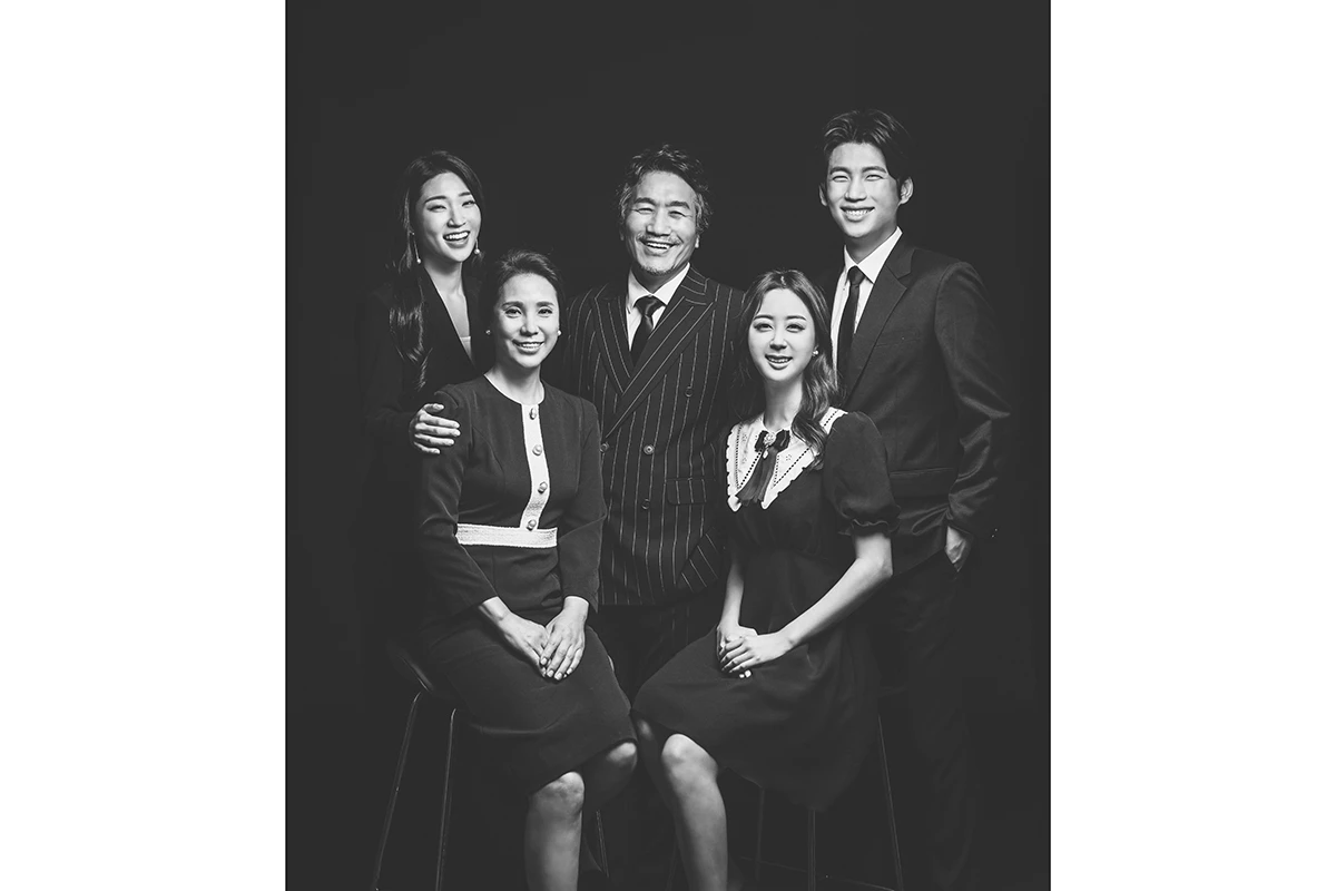 밝은 흰색 스튜디오에서 검정과 흰색 의상의 여러 세대 인물이 흰 배경과 나무 박스 주변에 앉고 선 사진이며, 나무 박스와 의자가 장면의 구성을 보완합니다.