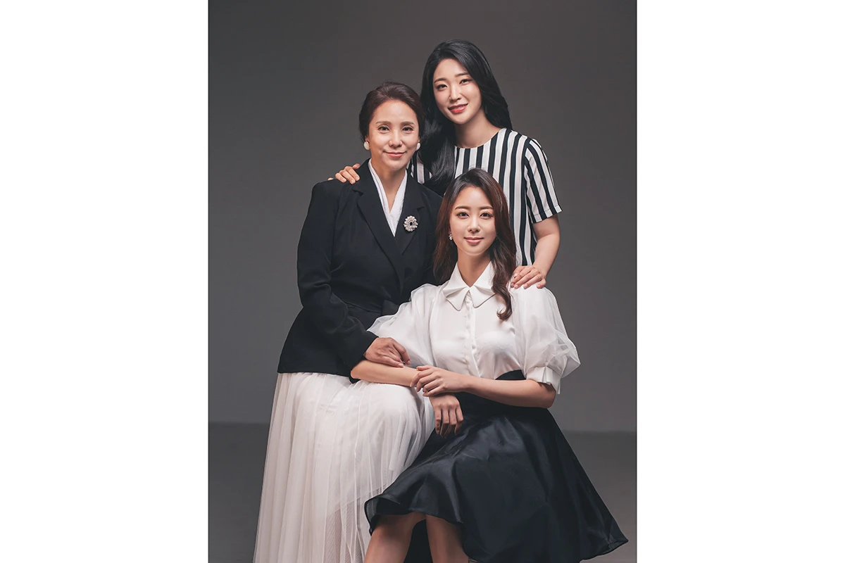 어두운 회색 스튜디오 배경에서 검정 의상의 성인 5명이 어두운 배경 앞에서 흑백 톤으로 촬영된 단체 사진이며, 의자와 흑백 처리가 장면의 구성을 보완합니다.