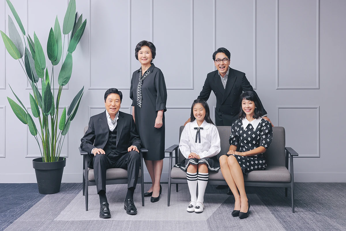 회색 스튜디오 배경에서 검정과 흰색 의상의 성인 5명이 선물 상자 앞에서 나란히 선 사진이며, 줄무늬와 체크 선물 상자가 장면의 구성을 보완합니다.
