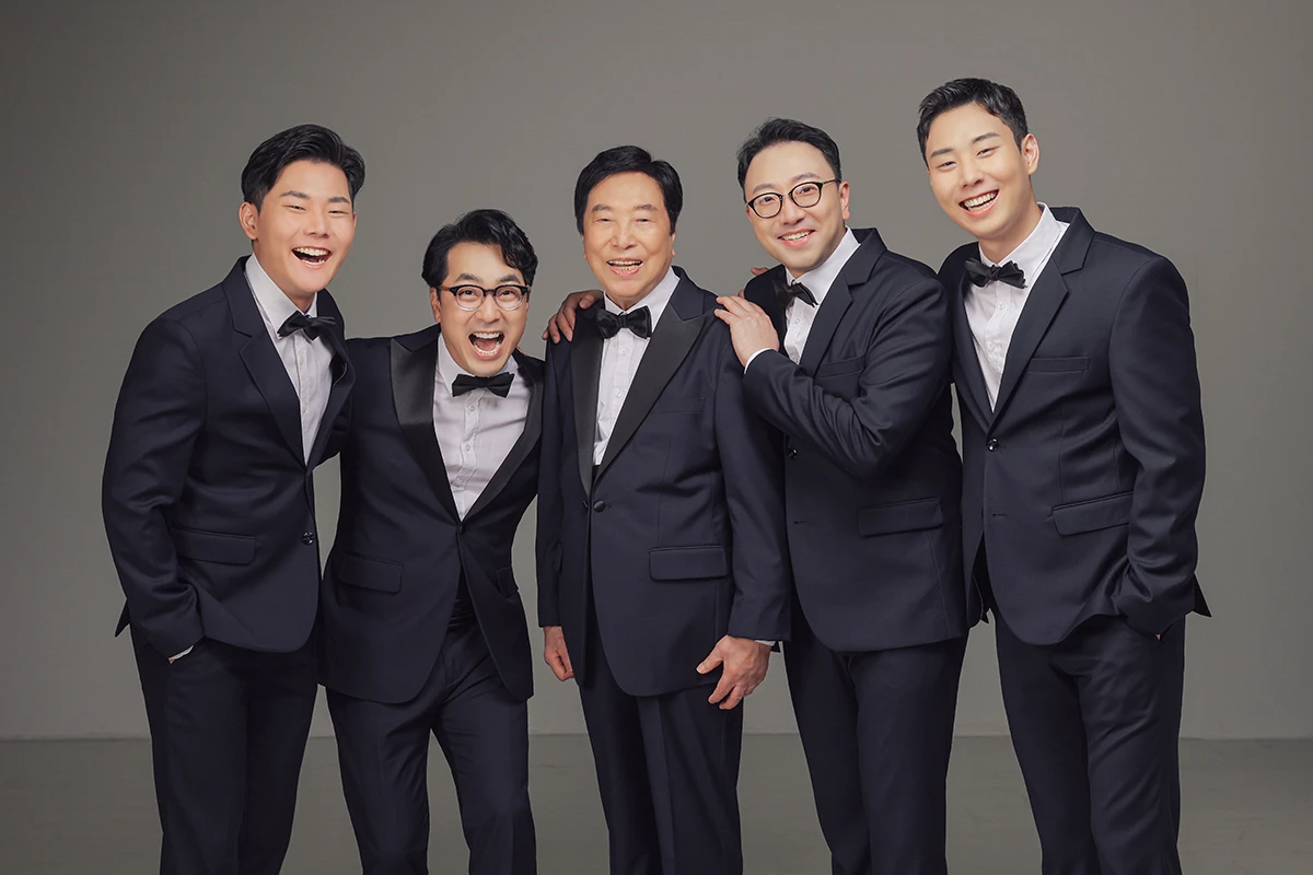 회색 배경과 검정 소파가 있는 스튜디오에서 검정 의상을 입은 성인 5명이 검정 소파와 회색 배경 앞에 배치된 사진이며, 검정 소파와 무늬 러그가 장면의 구성을 보완합니다.