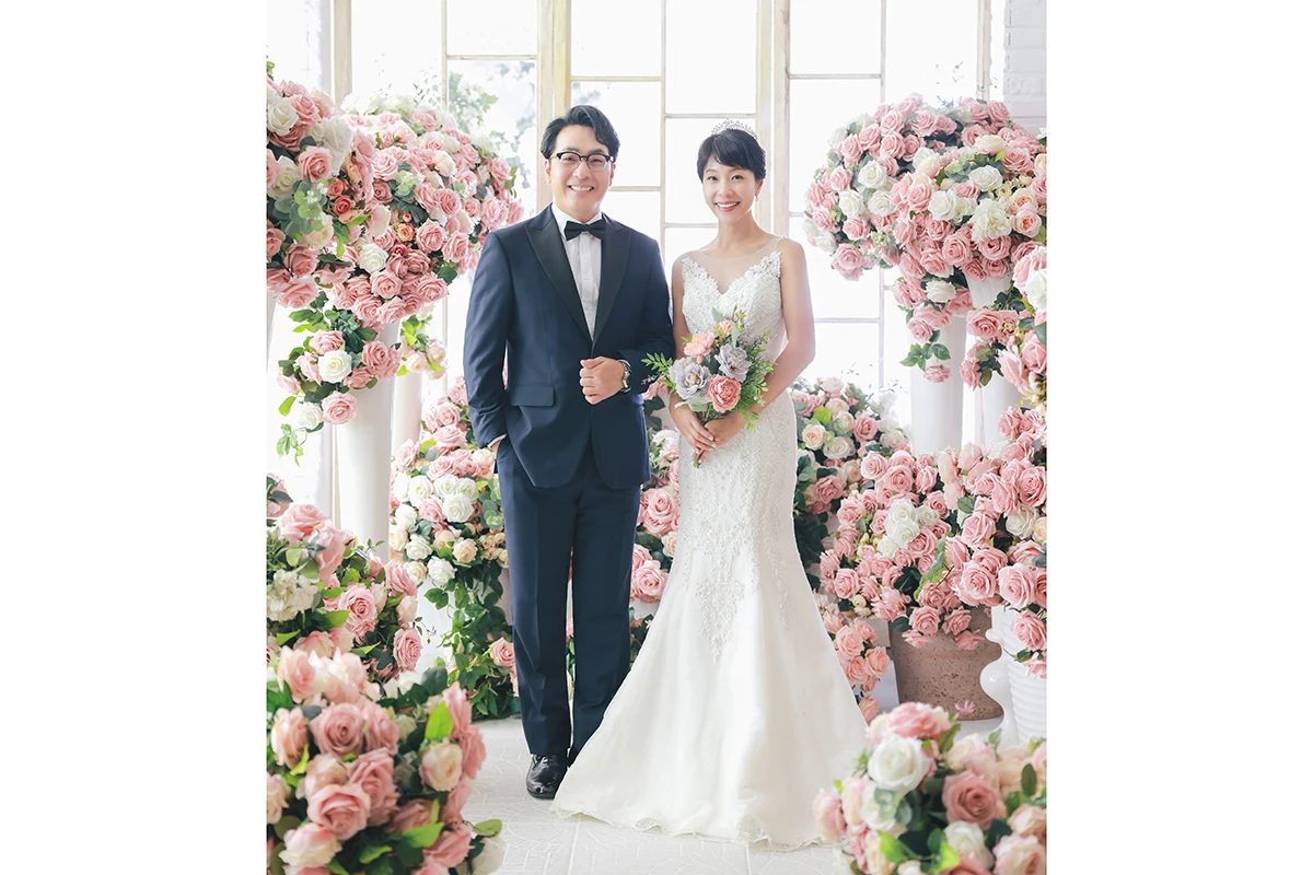 흰 벽돌과 큰 창이 있는 실내에서 화이트 드레스를 입은 성인 2명이 흰 벽돌과 창가 앞에서 서로 마주 보는 사진이며, 부케와 흰 꽃 장식가 장면의 구성을 보완합니다.