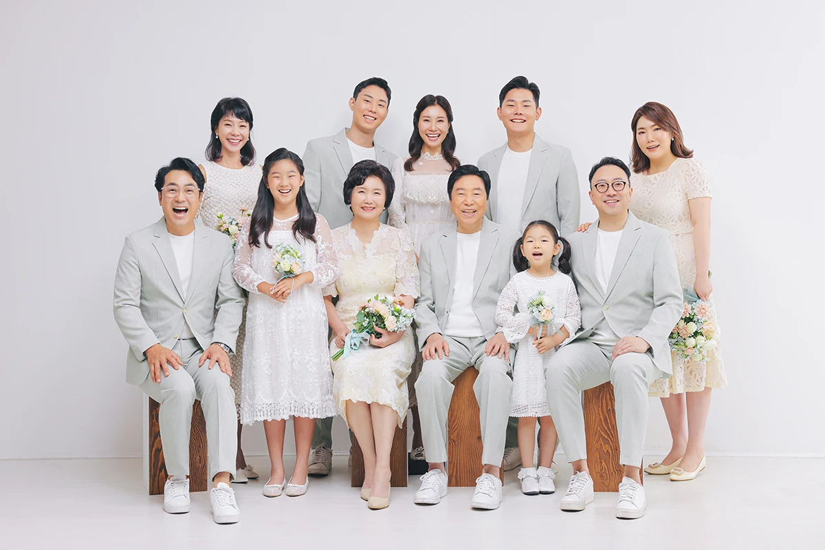 밝은 창가와 꽃 장식 세트에서 턱시도와 화이트 드레스를 입은 성인 2명이 꽃 장식 사이에 나란히 선 사진이며, 부케와 꽃 아치가 장면의 구성을 보완합니다.