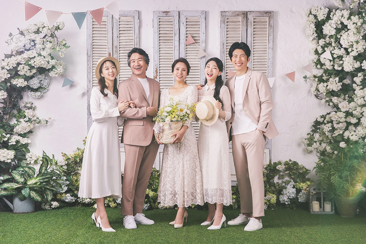 샹들리에와 소파가 있는 클래식 실내에서 화이트 드레스와 턱시도를 입은 여러 명이 클래식 실내 세트에 넓게 배치된 사진이며, 소파, 부케, 커튼가 장면의 구성을 보완합니다.