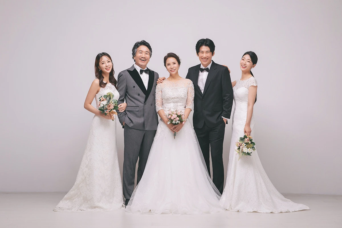 밝은 창가와 꽃 장식 실내에서 턱시도와 화이트 드레스를 입은 성인 3명이 꽃 장식과 창가 앞에 앉은 사진이며, 부케와 꽃 장식가 장면의 구성을 보완합니다.